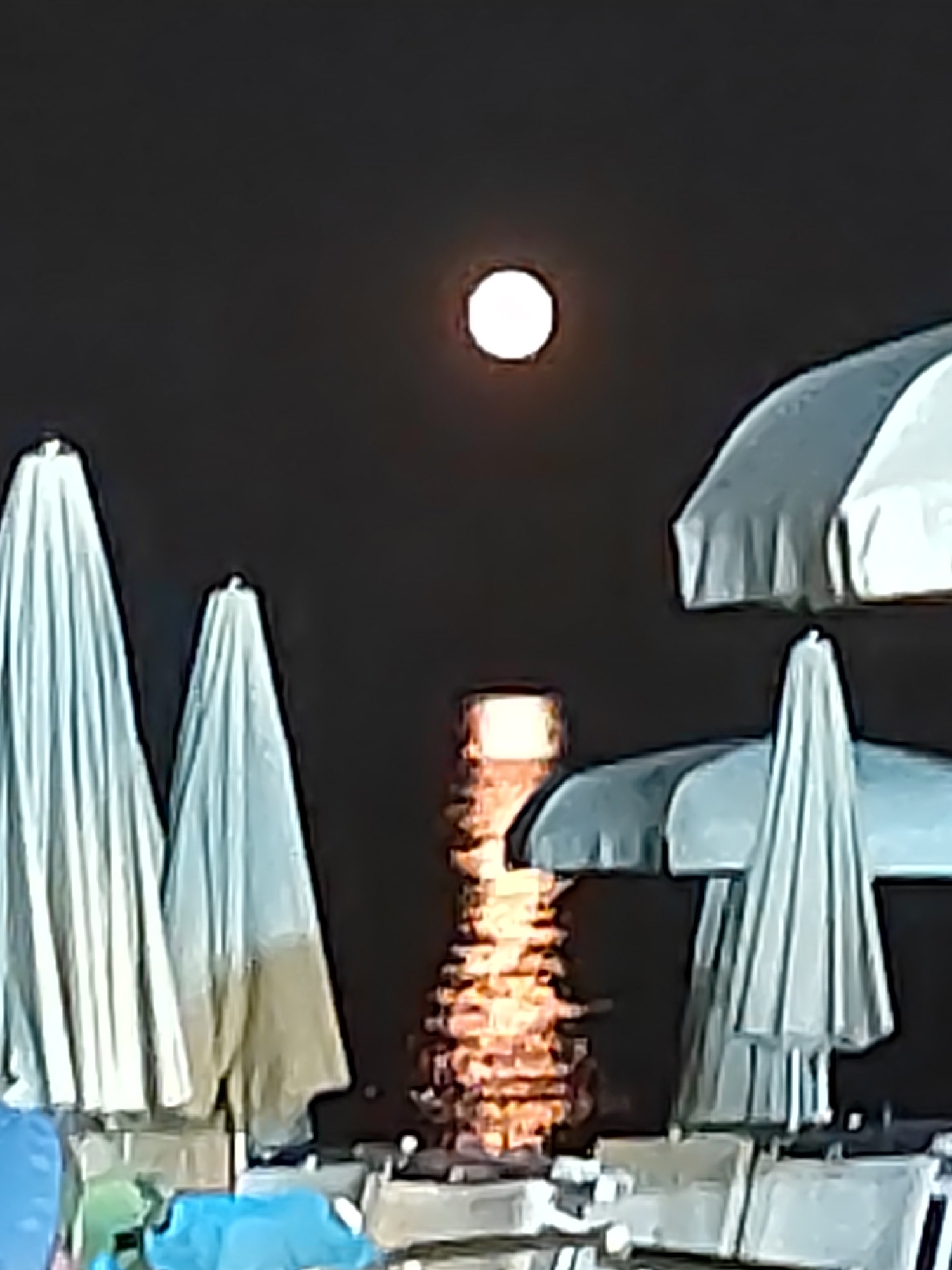 The image depicts a nighttime scene on a beach in Porto Recanati, Italy. The moon is visible as a bright, circular orb in the dark sky. In the foreground, several closed beach umbrellas are prominent, their fabric appearing light-colored. Behind them, the sea reflects the light from a distant source, creating a shimmering effect on the water's surface. Some beach chairs and tables are also visible in the lower part of the image, suggesting the presence of a beach establishment. The overall impression is a serene, nocturnal beach atmosphere.