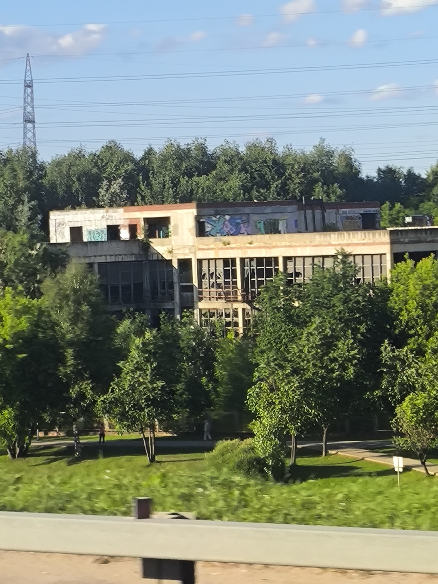 This media file captures an abandoned industrial building, possibly a factory or warehouse, surrounded by lush green trees and foliage. The building itself appears to be made of concrete and has a weathered, decaying facade with graffiti spray-painted on its upper level. Many of the windows are broken or missing, hinting at neglect and disuse.
In the foreground, a grassy area with some sparse trees and bushes suggests a park or landscaped area around the industrial structure. There are a couple of indistinct figures walking in the distance on a paved path, adding a sense of scale and human presence to the otherwise deserted scene.
The sky above is a clear blue with a few wispy white clouds, indicating it's likely daytime, perhaps late afternoon given the quality of light. The overall mood of the image is one of abandonment and decay, juxtaposed with the vibrant, persistent life of the surrounding nature. There are no discernible text elements in the image that provide specific location information or context. A tall transmission tower and power lines are visible in the background, suggesting the area is connected to utilities, despite the derelict state of the building.
