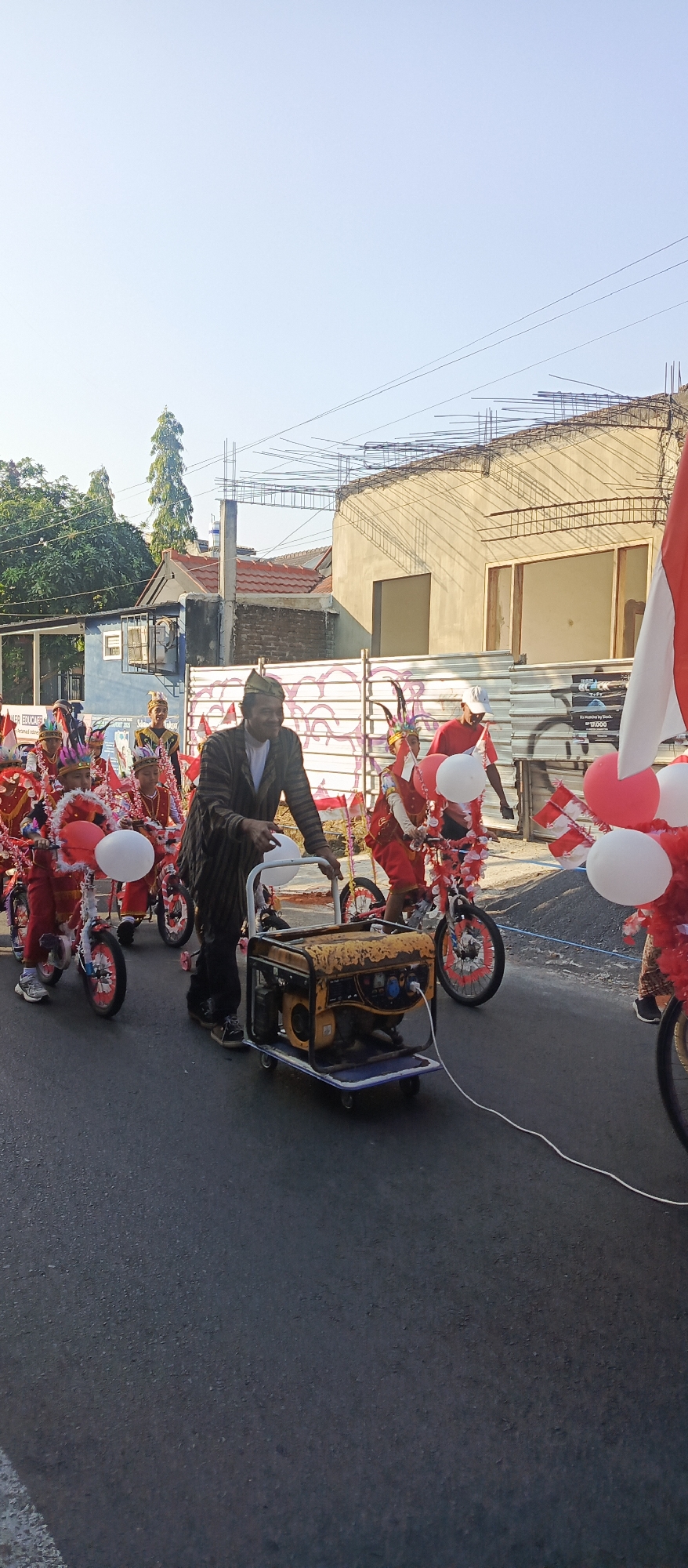 In Sekarpuro, Indonesia, a man in a patterned shirt and dark pants pushes a yellow generator on a cart. He is participating in a parade with several children dressed in red and white costumes, adorned with feathers and some holding balloons. The children are riding small, decorated bicycles. The parade is taking place on a paved road, with buildings and construction visible in the background. The sky is clear and blue.