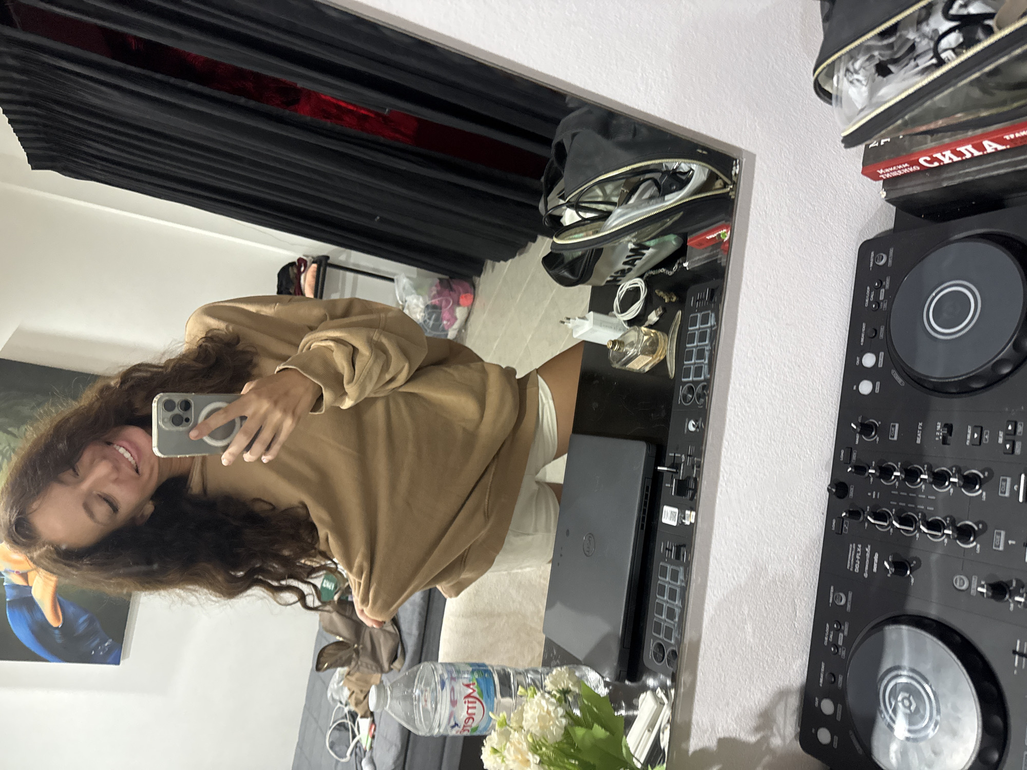 The image displays a mirror selfie of a young woman smiling and holding a light-colored iPhone, distinguishable by a visible MagSafe ring. She has long, wavy dark hair and is dressed in a loose-fitting tan hoodie and light-colored shorts.
The reflection reveals an indoor setting in Baan Chaweng, Thailand. Behind the woman, a grey sofa or bed is visible, along with a painting on the wall featuring blue and orange abstract shapes. A clear plastic bag containing clothes is also reflected.
The mirror rests above a dark surface or shelf, cluttered with various items. These include a closed black Dell laptop, a clear plastic bottle of Minera water, and a small bouquet of white flowers. Further along are a gold-toned perfume bottle, a white power adapter, a white charging cable, and a black toiletry bag with contents like eyeglasses. A black Pioneer DDJ-FLX4 DJ controller, labeled with "serato," dominates the right side of this surface.
Behind the mirror setup, black horizontal blinds or curtains are visible, with a hint of red fabric behind them. A stack of books includes one with a red spine and Cyrillic text that reads "МАКСИМ ГОРЬКИЙ СИЛА" (Maxim Gorky, Power/Strength). The overall impression is a casual and personalized indoor space. The woman's expression conveys happiness or contentment.
