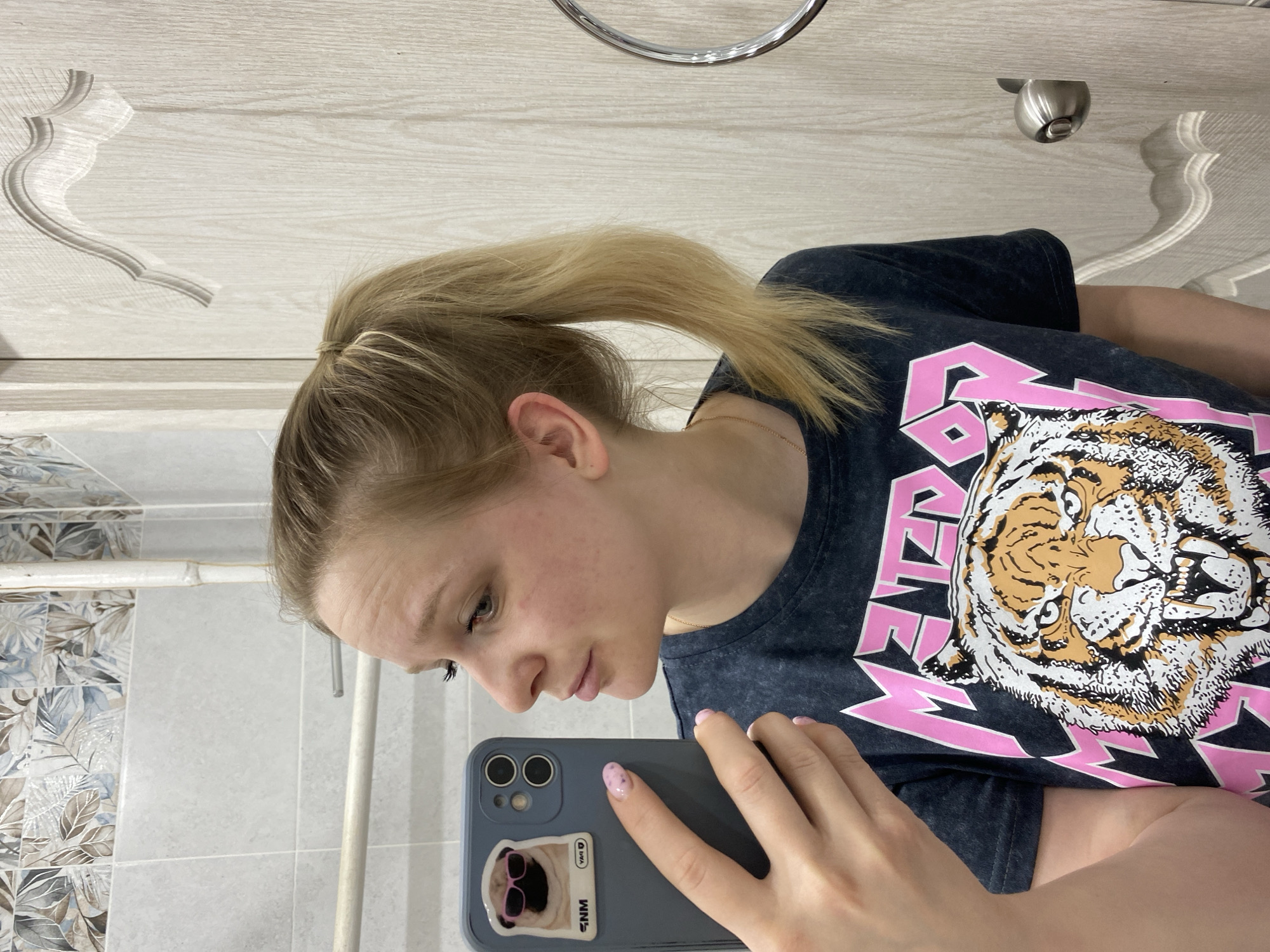 A young Caucasian woman with blonde hair pulled back in a ponytail is taking a selfie in what appears to be a bathroom. The bathroom has light-colored wood paneling on the upper walls, a chrome towel ring, and a patterned tile backsplash. The woman is wearing a dark T-shirt with a large tiger graphic and pink lightning bolt design. She is holding a blue smartphone with a sticker of a dog wearing sunglasses on the back. The location is Chaikovsky, Russia.