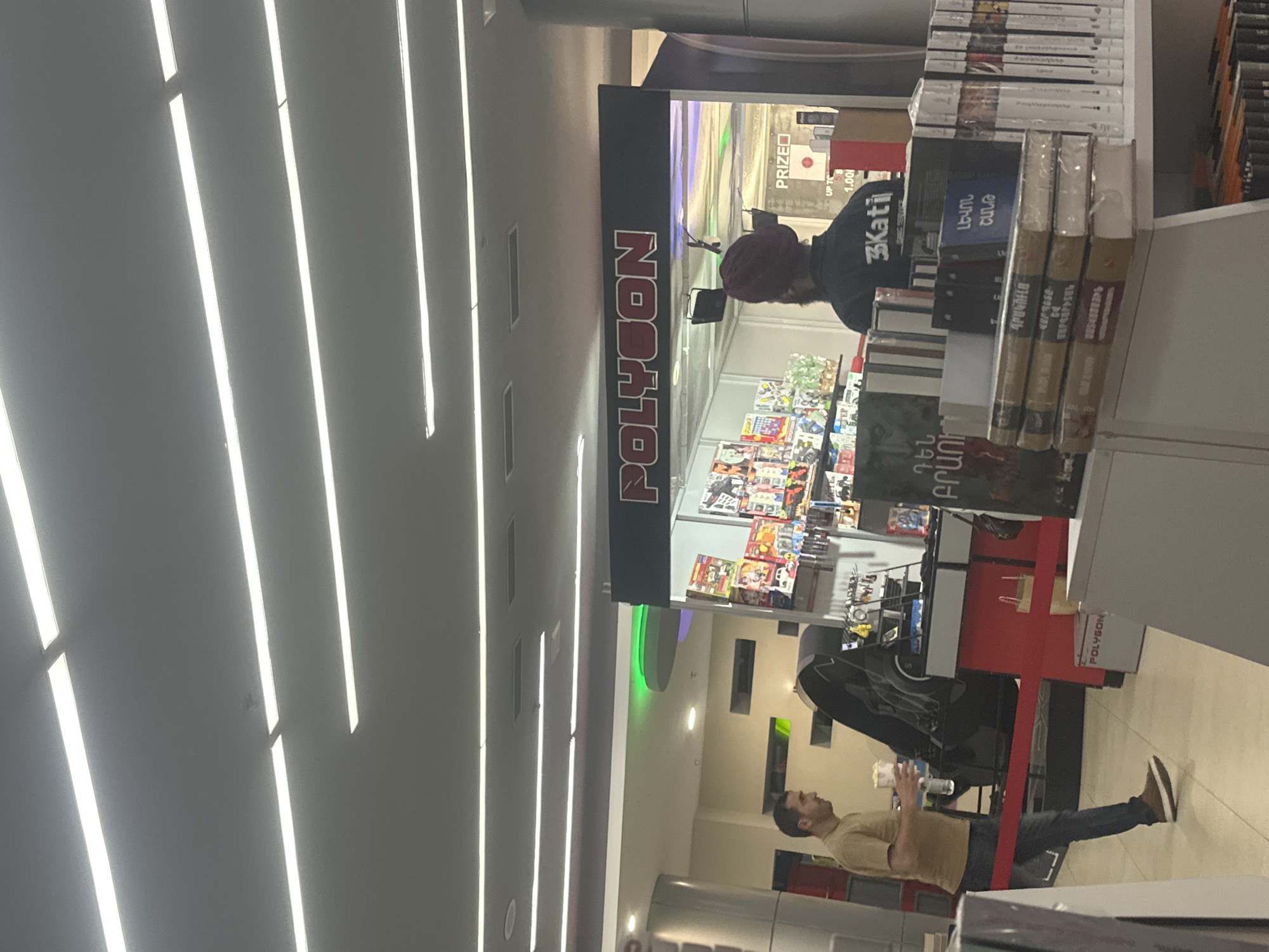 An indoor scene, likely within a retail establishment such as a bookstore, gaming store, or a section of a mall, is captured. The image is oriented vertically, showing a bright, well-lit environment with prominent linear fluorescent ceiling lights.
In the mid-ground, a man with dark hair and a beard stands facing left, wearing a light brown t-shirt and blue jeans with brown shoes. He holds a clear plastic cup in his right hand. To his right, a red and black booth or stall dominates the central area. The stall features a large black sign on top with the word "POLYGON" in bold, white, stylized letters. A smaller sign near the bottom of the stall also displays "POLYGON".
Behind the stall's counter, partially obscured, another person is visible from the back, wearing a dark top that appears to have "3Kati" written on it, and a dark red or maroon beanie. They seem to be standing behind a display of merchandise.
The stall itself and adjacent shelves are filled with numerous books and packaged items. Many book spines display text in Armenian script, including titles such as "ՆՎԵՐՆԵՐ" (Nverner - Gifts), "ԼԵԶՎԻ ՇԱՐԺԻՆ" (Lezvi Sharzhim - Language Movement/Engine), and "ԳՐԱԿԱՆՈՒԹՅՈՒՆ" (Grakanutyun - Literature). In the background of the stall, another sign is partially visible with "PRIZED" and "1.000" written on it.
The overall atmosphere suggests a casual shopping or browsing experience. The lighting is bright and even, indicating an indoor commercial space. No specific time of day or outdoor weather is discernible.