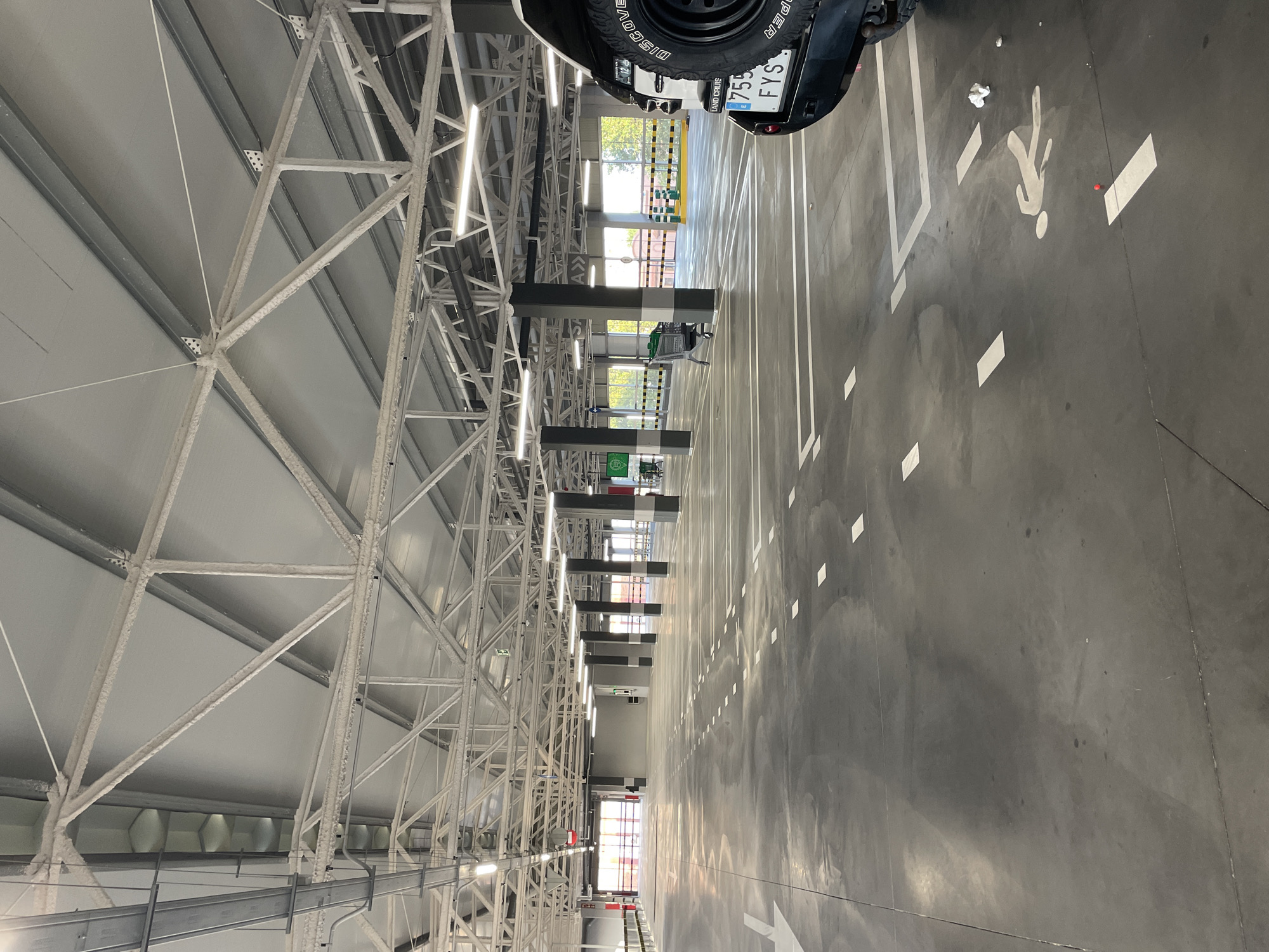 This is an image of an indoor parking garage. The garage is made of concrete and has metal beams and panels for its ceiling. The floor is also concrete, marked with white lines to designate parking spaces and a pedestrian walkway symbol. There are support pillars throughout the garage. The garage is mostly empty, with only a few shopping carts and the rear of a white SUV visible. The SUV has a license plate that reads "755 FYS". The scene appears to be clean and well-lit by overhead fluorescent lights. The location is La Línea de la Concepción, Spain.