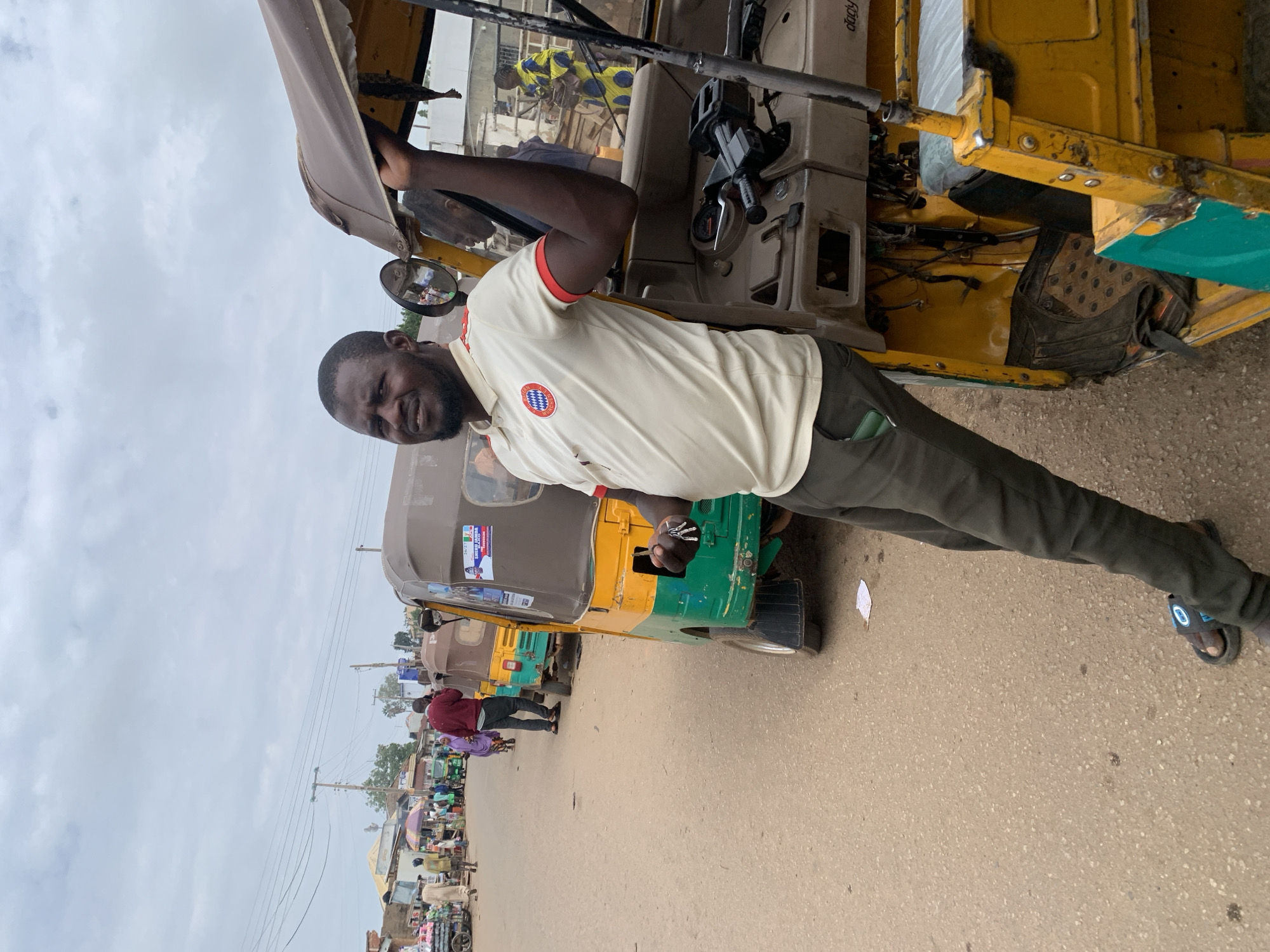 The image depicts a street scene in Gura Top, Nigeria, under an overcast sky. A dark-skinned man with a beard, wearing a cream polo shirt with red trim and a circular emblem (resembling a Bayern Munich logo), dark trousers, and black flip-flops, stands in the foreground. He leans against a yellow and green three-wheeled auto-rickshaw, holding a set of keys in his right hand, and looks towards the camera with a slight smile. The vehicle features a tan canvas roof and a dashboard displaying "dady".
In the background, the unpaved road is lined with several similar three-wheeled vehicles, some yellow and green, others brown. One vehicle prominently displays a political campaign poster with "SHEHU JANIDA MAKO". Numerous people are visible along the street; a man in a red sweater walks away, engaged in a phone call, followed by a woman in purple attire. Further back, market stalls with umbrellas and various goods line the street, with single-story buildings and utility poles under power lines completing the urban landscape.