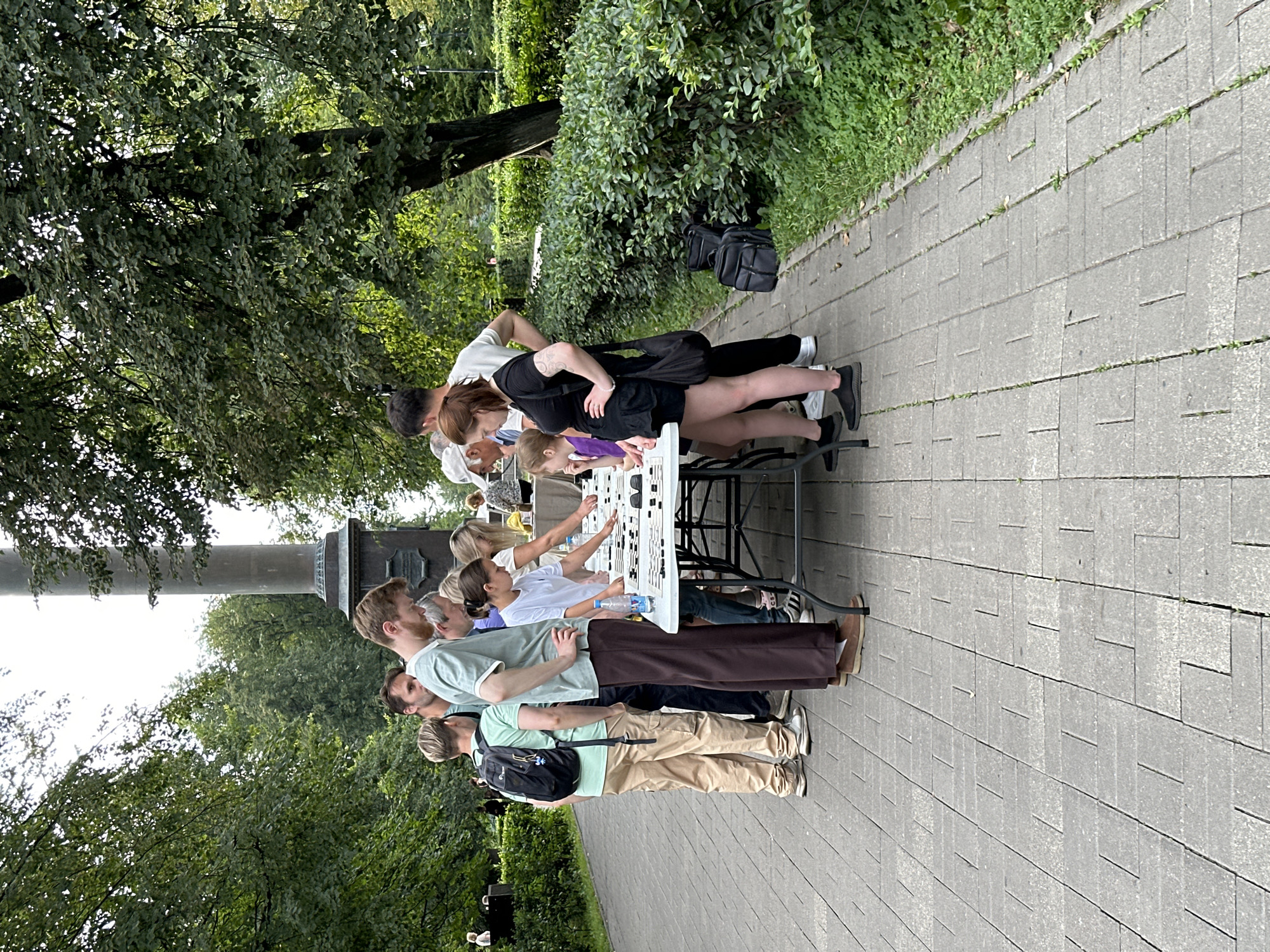 The image shows a group of people gathered around a table outdoors, possibly in a park. The table is covered with small items, and several individuals appear to be examining them. The background features trees, a tall monument or column, and a paved walkway. The people are casually dressed. There are no visible signs or indications that this scene is located in the city of Putyatino (Semivragovskaya S/A), Russia.