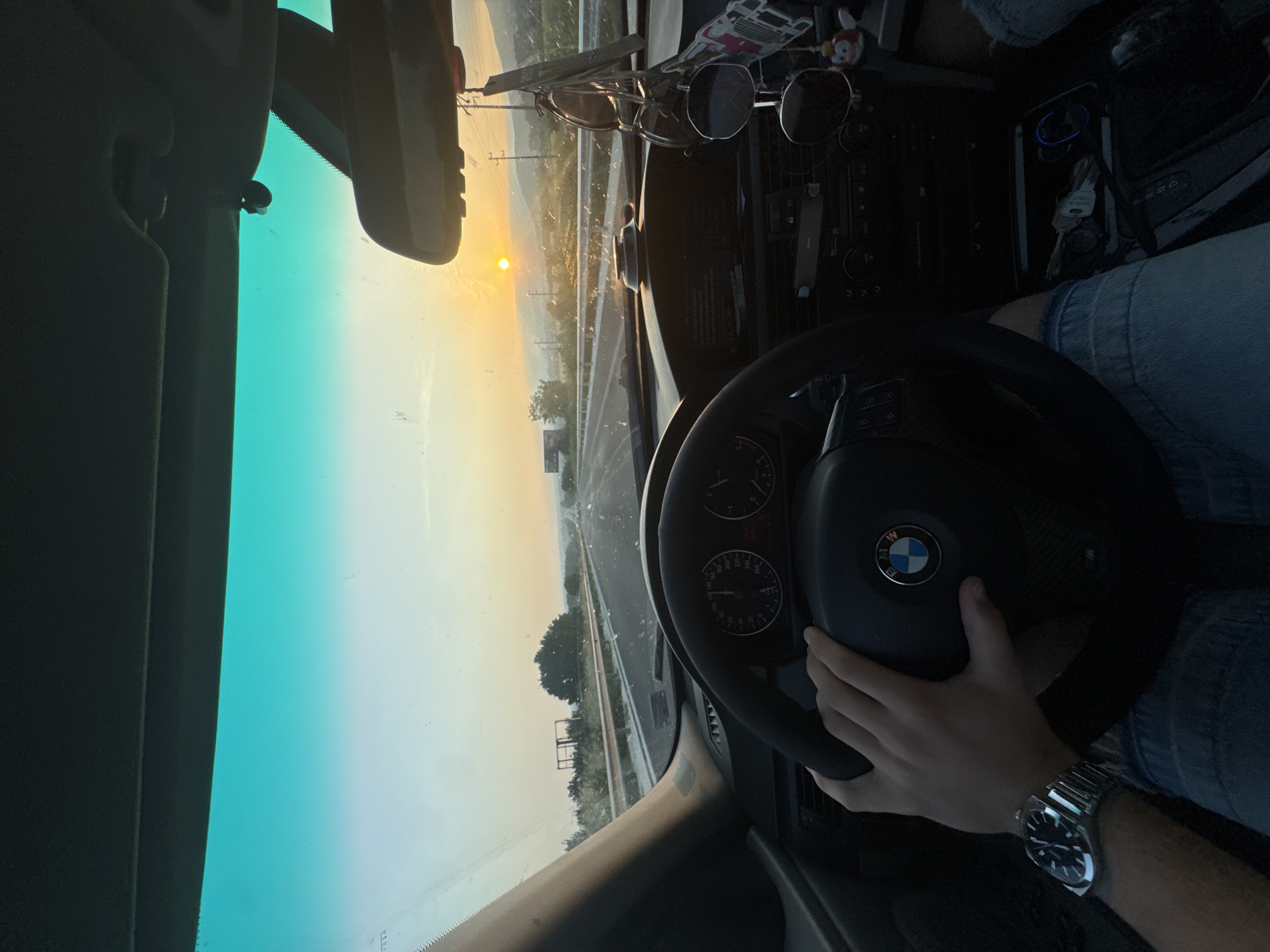 The image depicts a first-person perspective from inside a moving vehicle, specifically a BMW, traveling on a multi-lane highway at sunset in the vicinity of Ignatievo, Bulgaria.
The driver's right hand is positioned on the black, three-spoke steering wheel, which prominently features the blue and white BMW logo at its center. A metallic watch with a dark face is visible on the driver's left wrist. Below the steering wheel, the driver's legs are visible, clad in blue denim jeans.
The car's interior features a dark dashboard, with visible instrument gauges including a speedometer marked up to 260 km/h and a tachometer. The center console below the dashboard contains climate controls, an entertainment unit, and a small display screen. Keys are present in an open compartment near the gear shifter. A mobile phone appears to be placed in a holder on the dashboard.
The windshield is partially obscured by numerous small spots or smudges, indicating dirt. The upper portion of the windshield exhibits a distinct teal-colored tint. A rearview mirror is centrally positioned, with multiple pairs of sunglasses and various decorative charms hanging from it.
Beyond the windshield, the highway extends into the distance under a clear, warm-toned sky. The sun, a bright yellow orb, is low on the horizon, slightly to the right of the center frame, indicating sunset. The landscape consists of a highway with guardrails on both sides, bordered by green vegetation including trees and shrubs. In the background, distant mountains or hills are visible. Utility poles and power lines traverse the scene on the right side. An empty billboard frame is situated on the left side of the highway, while another large sign structure is dimly visible further down the road on the right.