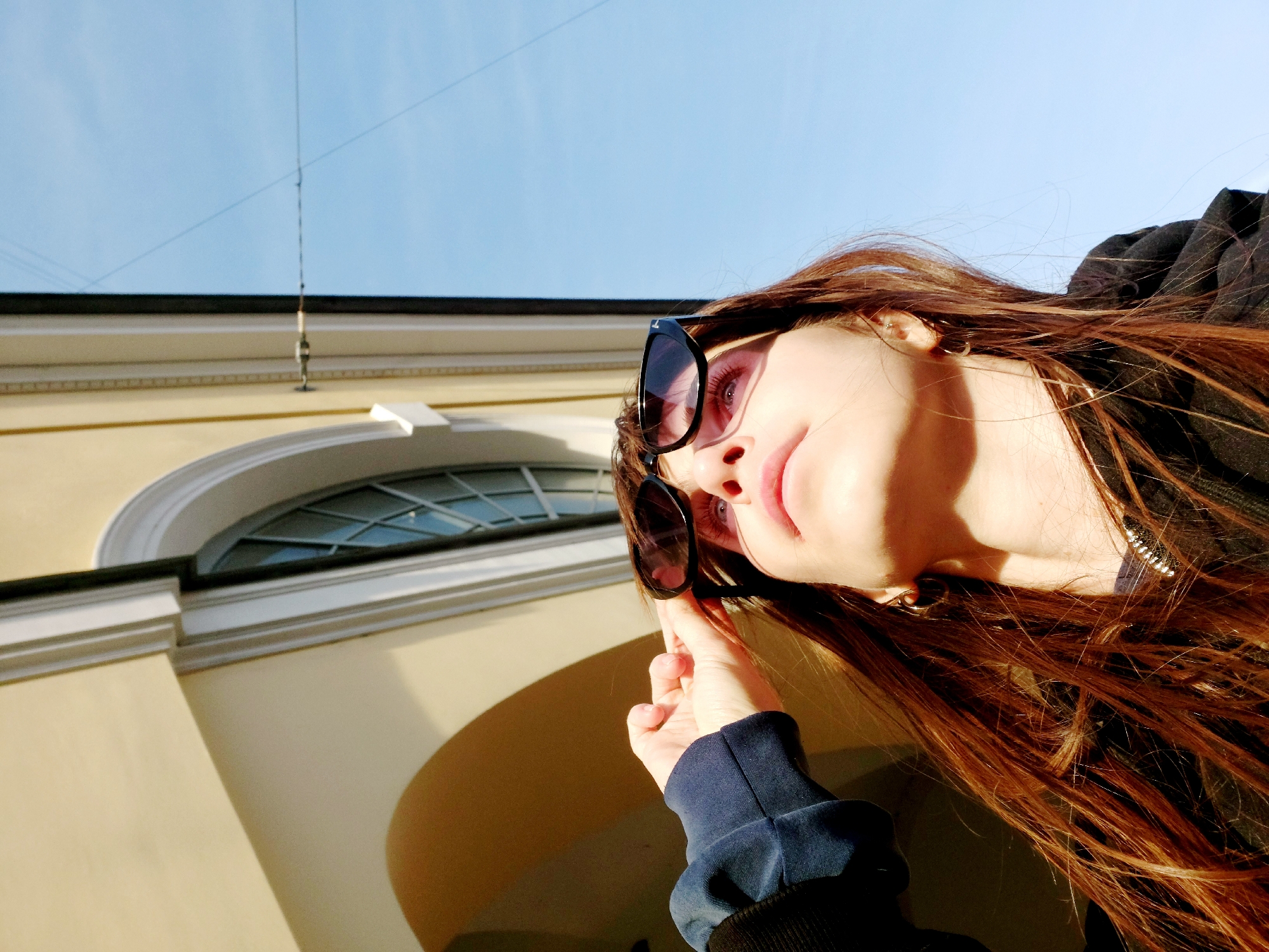 A low-angle, eye-level shot depicts a young woman wearing sunglasses, set against a backdrop of a pale yellow building and a clear blue sky. The woman is angled slightly toward the viewer, with her face and long, reddish-brown hair prominently displayed. Her right hand is visible, holding the sunglasses near her face. The building, partially visible, features a large arched window with a grid pattern. A thin, dark wire stretches across the sky in the background.
Based on the architectural style of the building, particularly the window and color, and the woman's appearance, the location is very likely in Saint Petersburg, Russia, although this determination is based only on the visual content of the image. The photograph appears to be taken outdoors during daylight hours, with sunlight illuminating the scene.