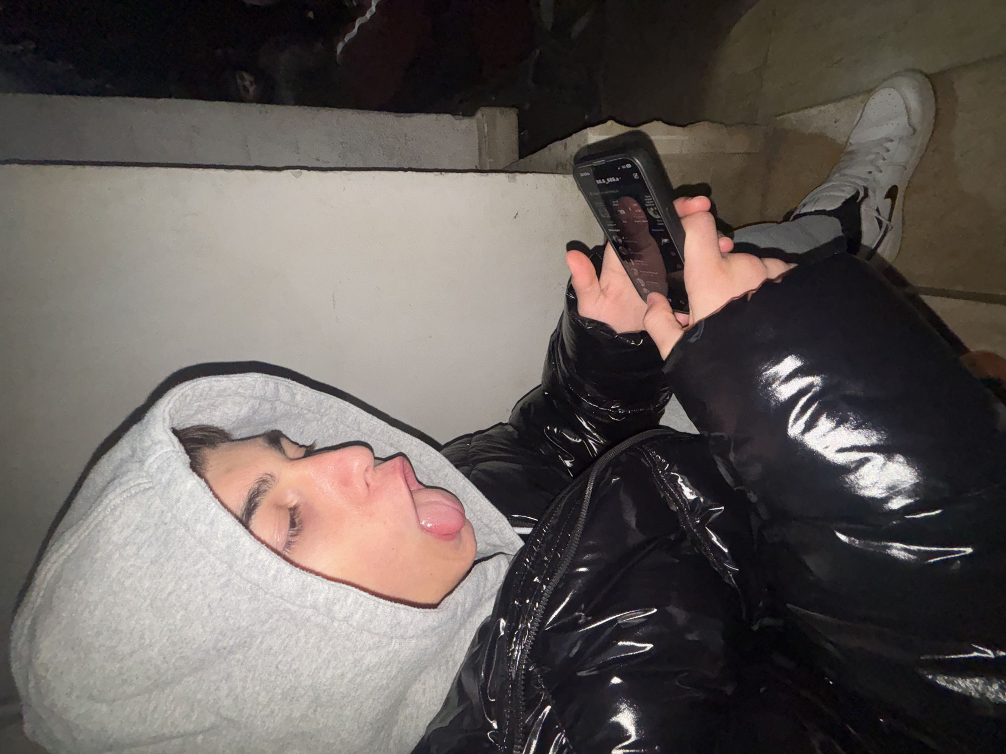 A young person in a grey hooded sweatshirt and black puffer jacket lies at an angle on a light-colored concrete or stone structure, possibly steps. They are holding a smartphone in their hands and looking at the screen. Their tongue is sticking out. A white sneaker is visible in the upper right. The location is Lehchevo, Bulgaria. The ambient lighting suggests it is nighttime.