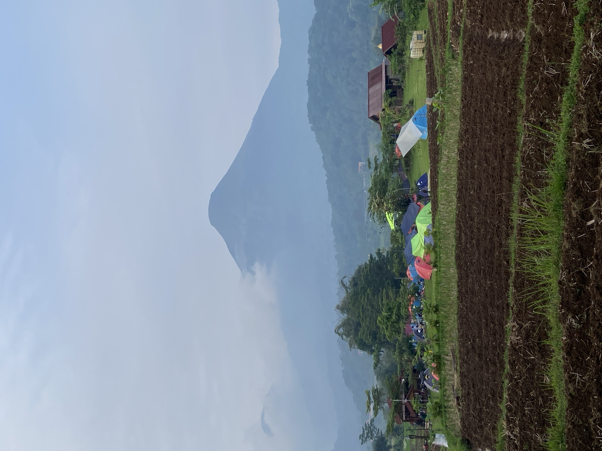 The image captures a campsite in Tamiajeng, Indonesia, bordered by terraced rice paddies. Various tents, mostly bright in color, are clustered among trees. In the background, a large, hazy mountain dominates the landscape. A few buildings are visible amidst the trees near the campsite. The overall atmosphere suggests a misty or overcast day.