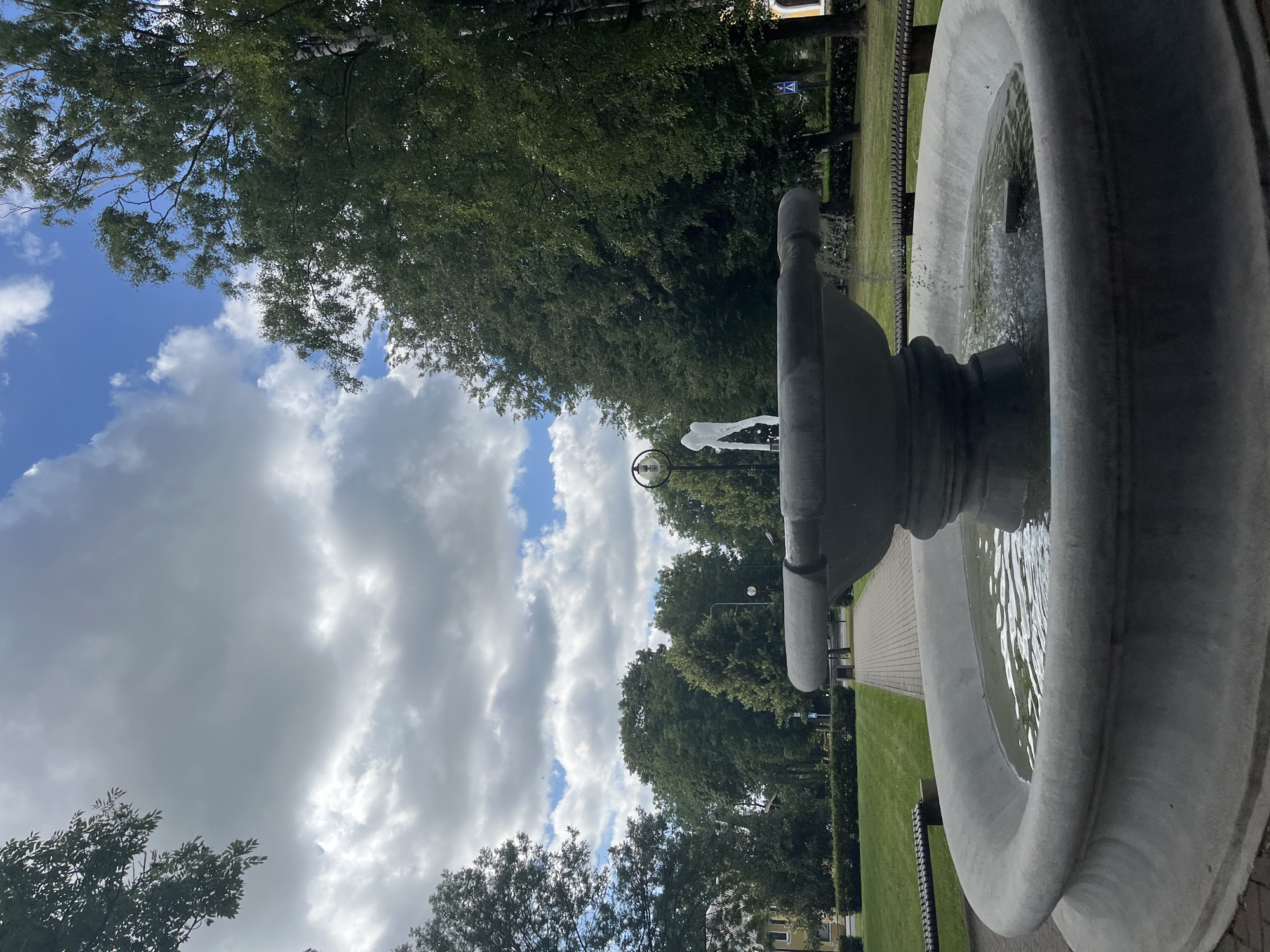 A low-angle, full shot shows a large, circular, concrete fountain in a park setting. The fountain's basin is filled with water, and a thin stream of water arcs upwards from the center. The fountain is made of weathered gray concrete, with a stepped base and a wide rim. Surrounding the fountain is a paved walkway, and beyond that, a well-maintained green lawn. In the background, dense trees line the perimeter of the park. A few park benches are visible along the walkway. The sky above is a mix of blue patches and large, dramatic gray clouds. The overall impression is a tranquil, public park in Kohtla-Järve linn, Estonia.