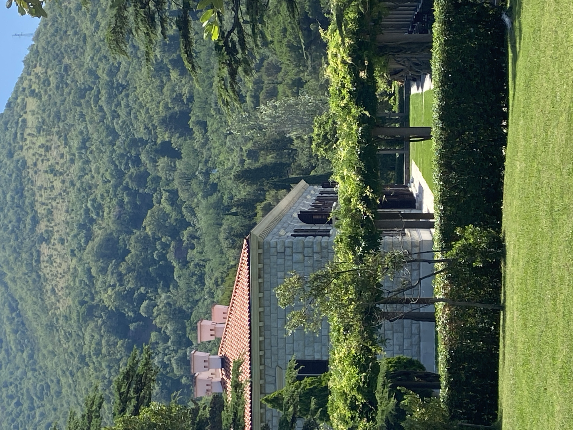 The image shows a large stone building with a red tiled roof and distinctive square chimneys, situated in the foreground of a lush green mountainous landscape. The building features a colonnaded walkway with climbing vines creating a natural pergola. A manicured hedge separates the building's grounds from a well-maintained lawn. The backdrop is a steep, tree-covered mountain slope, indicative of a natural, possibly protected, area. The architecture and the mountainous setting suggest a location in the Mediterranean region. This scene is consistent with views found in and around Sveti Stefan, Montenegro, known for its historic island and surrounding mountainous terrain. No people or specific actions are visible. The environment is serene and natural, with a focus on architecture integrated into the landscape.