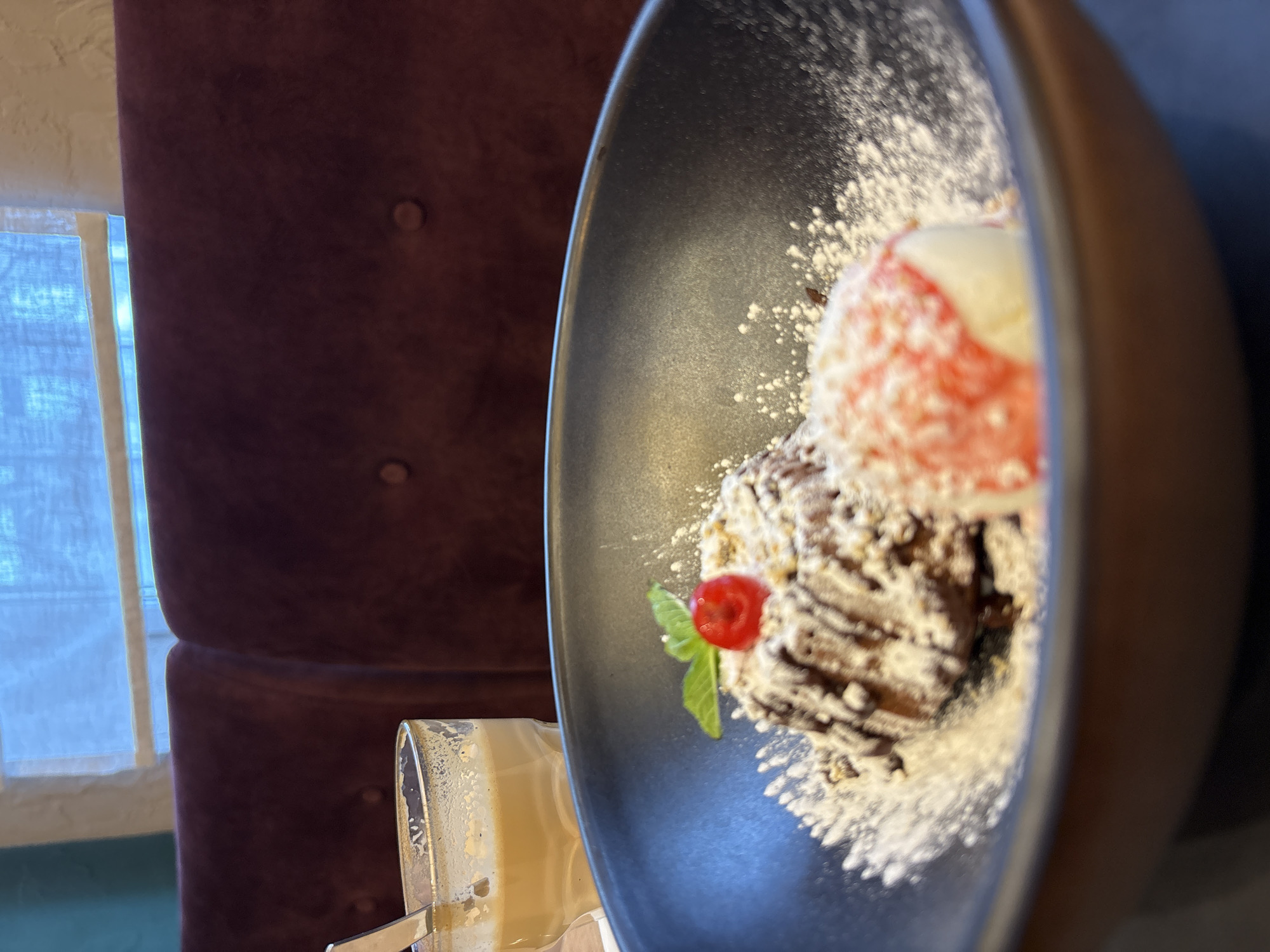 A close-up shot features a dark, possibly black, serving bowl filled with what appears to be a dessert. The dessert consists of a brownie or similar baked good heavily dusted with powdered sugar, topped with a scoop of ice cream that has a pink or red hue and a dollop of white topping. A small red fruit, possibly a cherry or berry, is positioned near a few green leaves as a garnish. A glass of a milky or creamy-looking beverage is partially visible at the bottom left of the frame.
The background includes a tufted burgundy chair and a window with a white frame, suggesting an indoor setting within a restaurant or cafe. There is no confirmed evidence of this photograph being taken in Neftekamsk, Russia.
