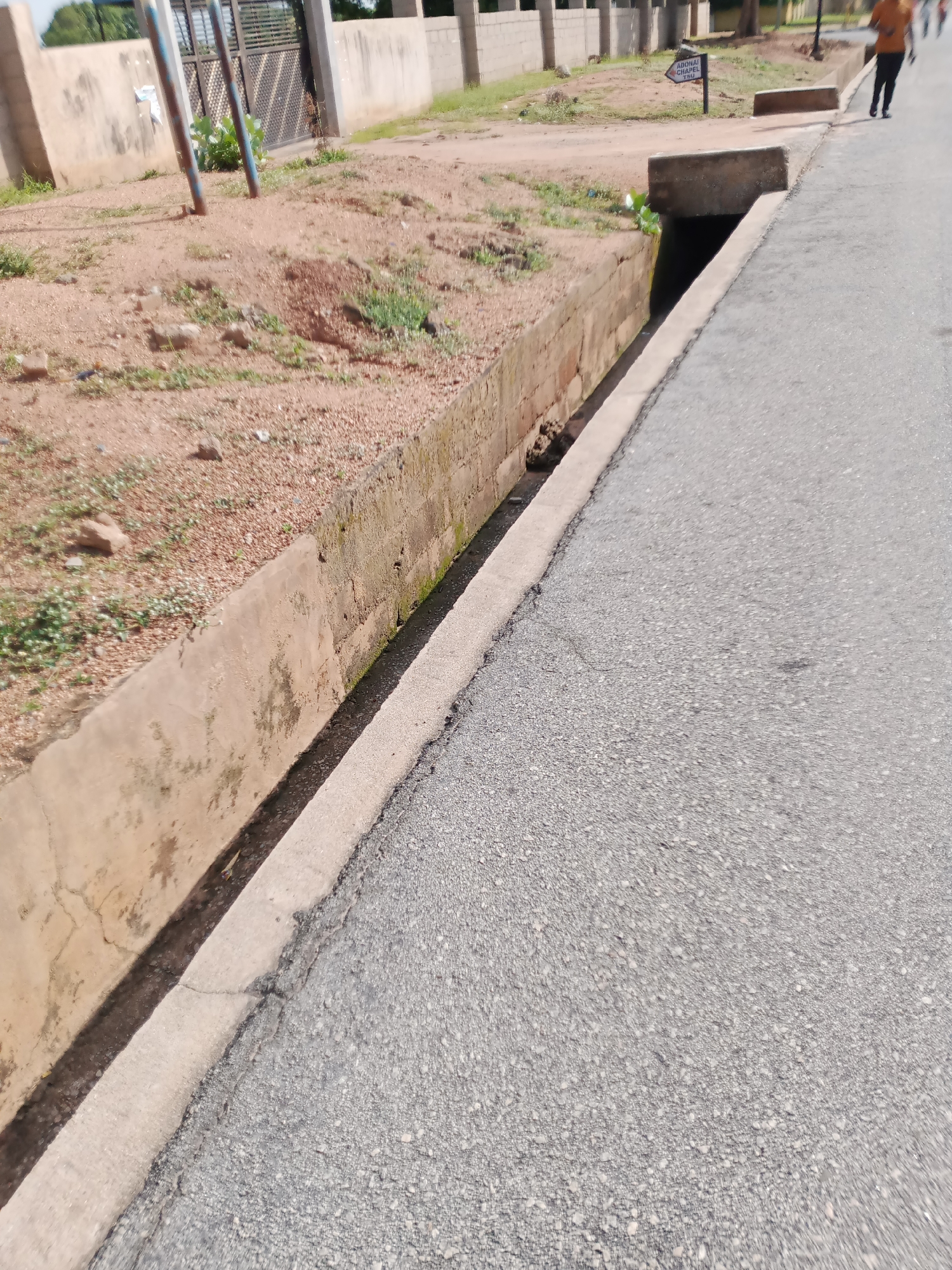 This is a ground-level, full-frame, eye-level shot of a concrete drainage ditch running alongside an asphalt road in Jalingo, Nigeria. The ditch is filled with dark water and debris. The road occupies the right two-thirds of the frame, showing a cracked asphalt surface. To the left of the road, a concrete wall borders the drainage ditch. Beyond the ditch, a dirt embankment with sparse vegetation and scattered rocks is visible. In the background, a fence and buildings can be seen. A sign pointing to the left reads "ADONAI CHARITY SCHOOL." A person in an orange shirt and black pants is walking away from the camera on the road, and another person is partially visible in the far background. The lighting suggests it is daytime.