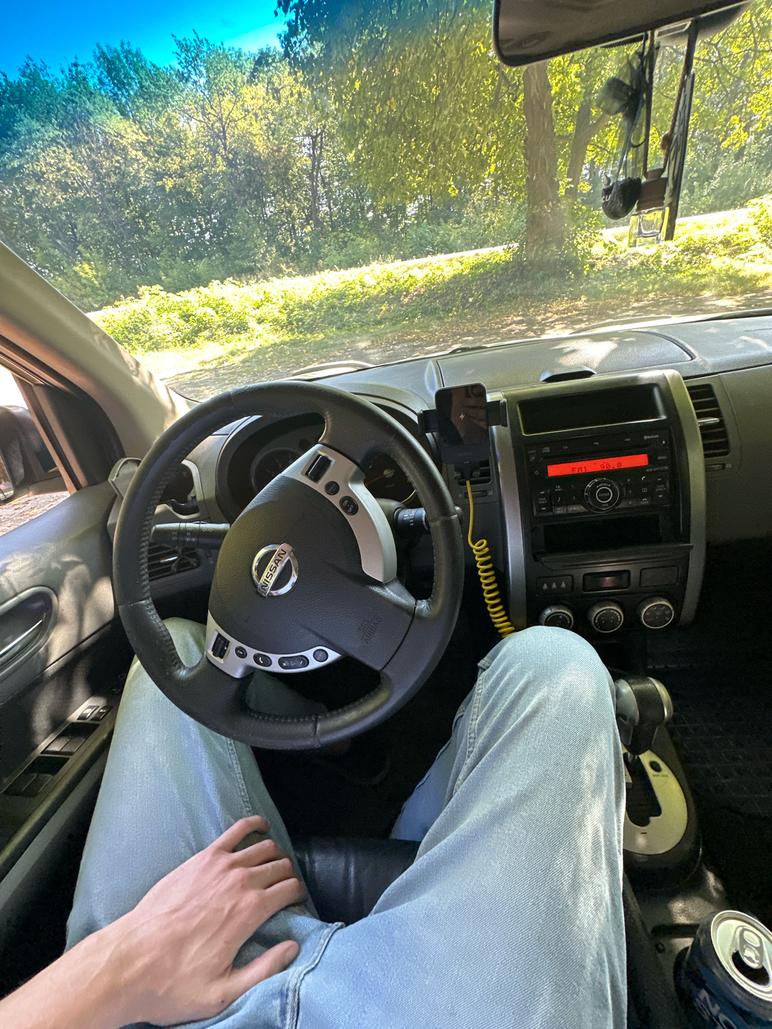 This is a first-person perspective shot from inside a car, showing the dashboard, steering wheel, and the driver's legs and hand. The view outside the windshield is of a tree-lined road with sunlight filtering through the leaves, suggesting a rural or suburban setting. The car's radio display shows "FM1 90.8". A can of soda is visible next to the gear shift. The image does not contain any information that would allow for its placement in Shatava, Ukraine, or any other specific city.