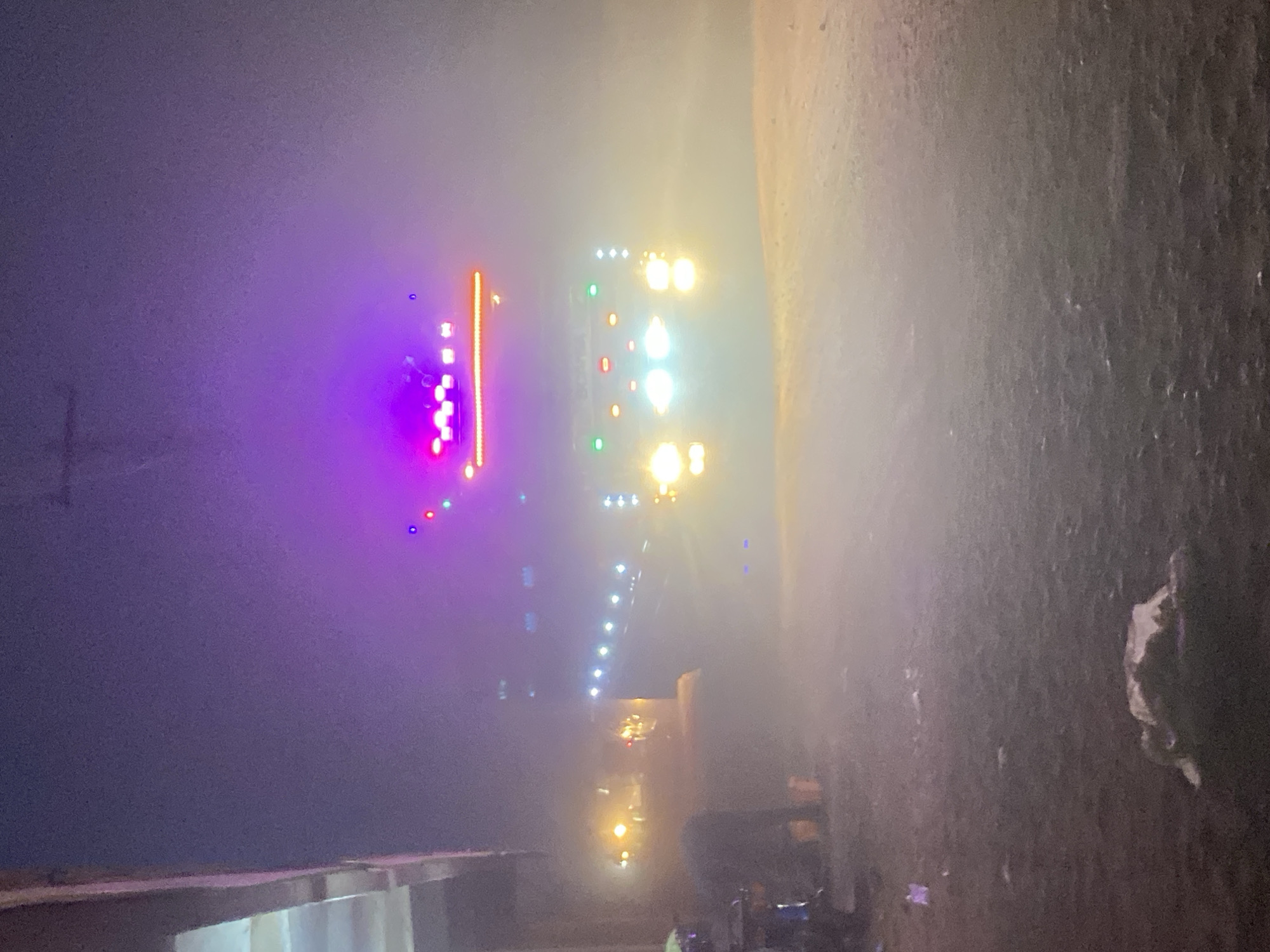 The image, likely taken in Tudun Wada, Nigeria, at night, shows a brightly lit truck or vehicle approaching on a street. The vehicle is adorned with multiple lights, including headlights, and purple and orange decorative lights. The environment is dark with limited visibility, possibly due to mist or fog. The foreground shows the side of a building or structure and some indistinct objects on the ground. There may be people in the scene, although they are difficult to discern.