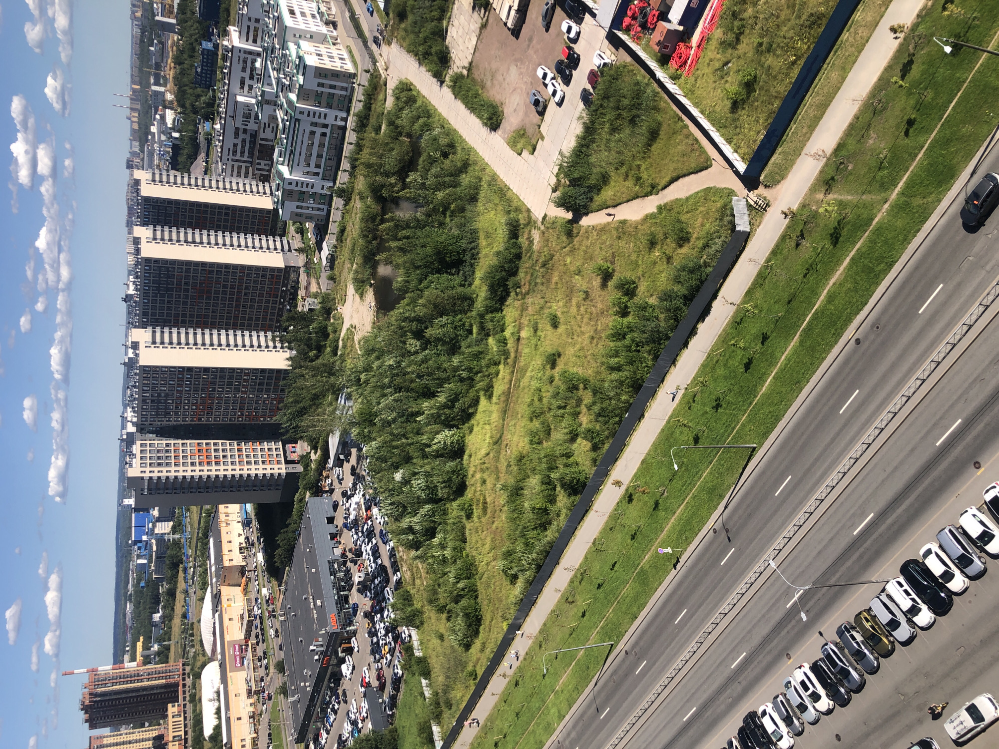 An aerial shot captures a contemporary urban landscape under a clear blue sky dotted with white clouds. The scene is dominated by a mix of residential buildings, commercial structures, and undeveloped green spaces. In the foreground, a multi-lane road runs diagonally across the frame, with a row of parked cars lining one side. To the left of the road, a grassy median strip is interspersed with young trees and lampposts.
Further into the image, a large, sprawling commercial complex with a dark roof and prominent "LADA" signage is visible, surrounded by a vast parking lot filled with vehicles. Beyond this, several tall, modern residential towers with beige and grey facades rise prominently, interspersed with clusters of dense green trees. Between the residential buildings and the commercial area, a small, dark pond reflects the surrounding foliage. To the right, a developing area shows white, multi-story buildings and construction materials, including bright red coiled pipes. The overall impression is one of urban expansion, with a balance of built environments and natural elements. A few individuals are seen walking on a paved path adjacent to the grassy areas.
