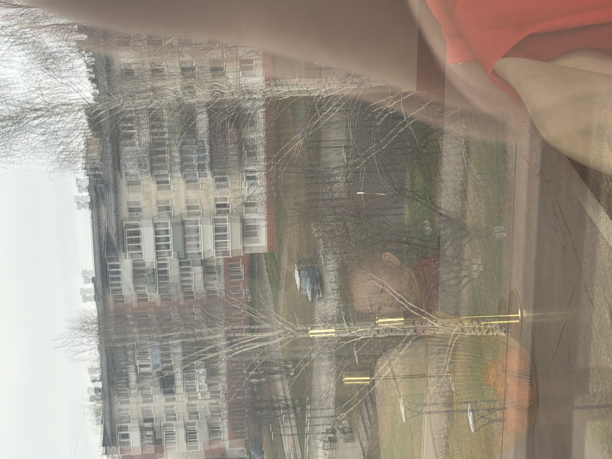 The image shows a view from inside a building, likely through a window. Outside, there is an apartment building, trees without leaves, a park area, a road and a parked vehicle. The location is likely in Chaikovsky, Russia, based on the urban architecture of the apartment building.
There are no clear people visible in the image, but there is a reflection of what appears to be a person (possibly a woman) taking the picture. The reflection shows a light-skinned person with light hair, wearing a beige top and an orange piece of clothing.
The overall environment is urban, with a mix of residential buildings and green spaces. The lighting suggests an overcast day.