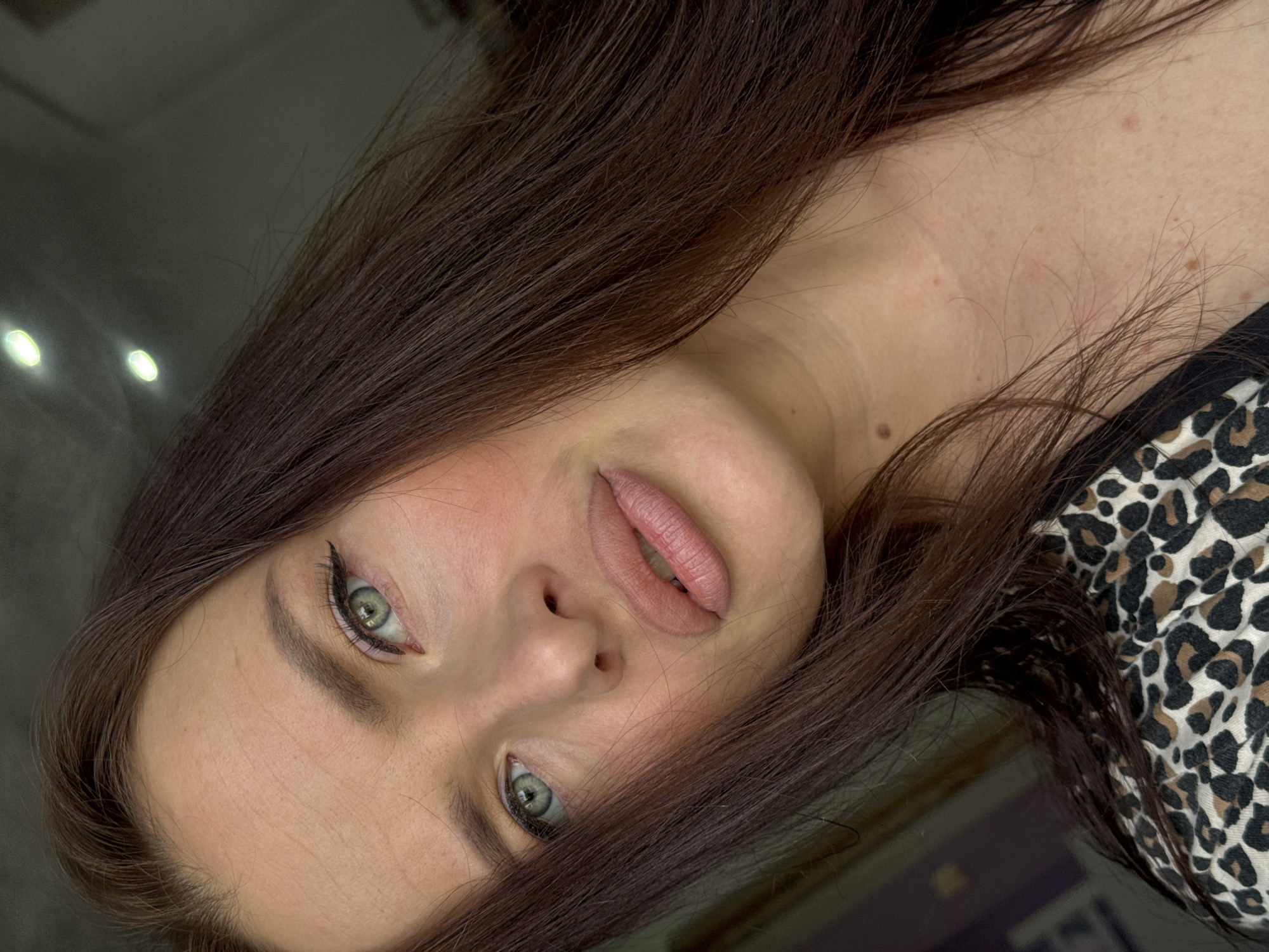 The image shows a woman's face close-up. She has fair skin, brown hair, and green eyes with eyeliner. Her lips are slightly parted. She is wearing a leopard print top. The background is dark and blurry with some light reflections. There is no indication of the location being Rusalevka, Russia.