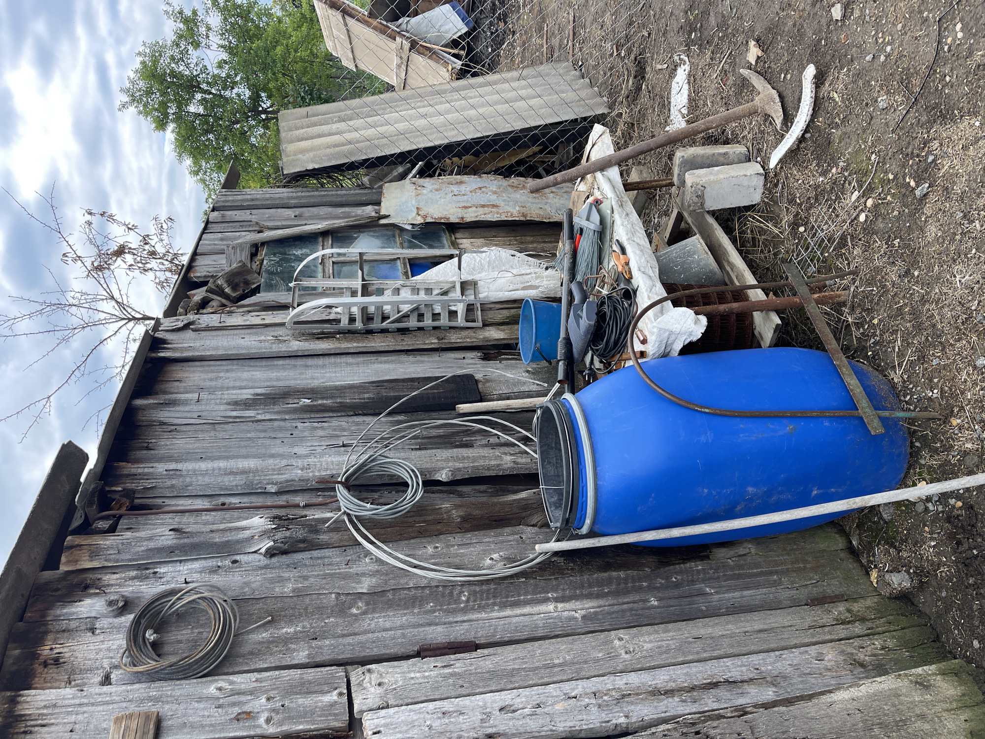 The image depicts a cluttered outdoor scene adjacent to a weathered wooden shed in Левокумка, Russia. A large blue plastic barrel stands prominently next to the shed. Several coils of wire are attached to the shed's exterior, along with a rusted metal object. In front of the shed, a makeshift table is covered with a white tarp, holding various tools including a drill, pliers, and a broom. A blue bucket and a piece of corrugated metal are also visible near the table. The ground is covered with dirt, dry grass, and debris. A pickaxe leans against the shed, and two concrete blocks are situated on the ground. In the background, a chain-link fence, some construction materials, and trees are present, suggesting an agricultural or storage area. The sky is overcast. There are no people or animals visible in the image.