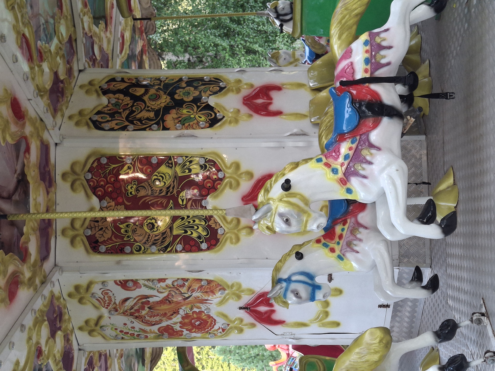 The image shows a close-up of a carousel, likely in Chaikovsky, Russia, with a focus on two white carousel horses. The horses are positioned side-by-side, facing right, with their bodies angled slightly towards the viewer. They are adorned with colorful saddles and decorative blankets. The horse in the foreground is predominantly white with a blue bridle and a saddle decorated with red, pink, blue, and purple geometric patterns. The horse behind it shares a similar design but is partially obscured. Both horses have black hooves and yellow supports beneath them.
The carousel structure is visible behind the horses, featuring panels decorated with traditional Russian folk art, specifically depicting stylized birds and floral motifs in red, yellow, black, and green. The carousel canopy is also visible at the top, with ornate golden and red designs. A yellow rope or pole runs vertically down the center of the carousel's interior. The environment appears to be outdoors, with lush green foliage and trees visible in the background, suggesting a park or amusement area. The ground beneath the carousel horses is a metallic, textured surface. The overall scene is brightly lit, indicating daytime.