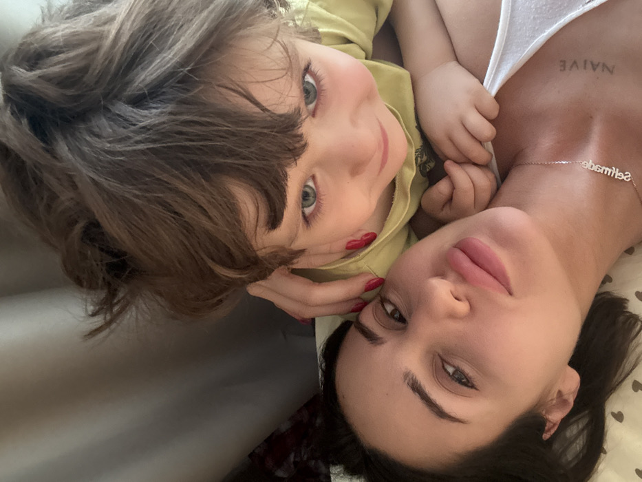A woman and a child pose closely together, likely for a selfie. The child, wearing a yellow shirt, has brown hair and striking blue-green eyes. The woman, with red nails, displays a tattoo reading "NAIVE" on her chest and a necklace spelling out "Seltmade." They are positioned against a soft, patterned surface, suggesting they might be lying on a bed. The image is taken from a slightly high angle, capturing both faces in close proximity.