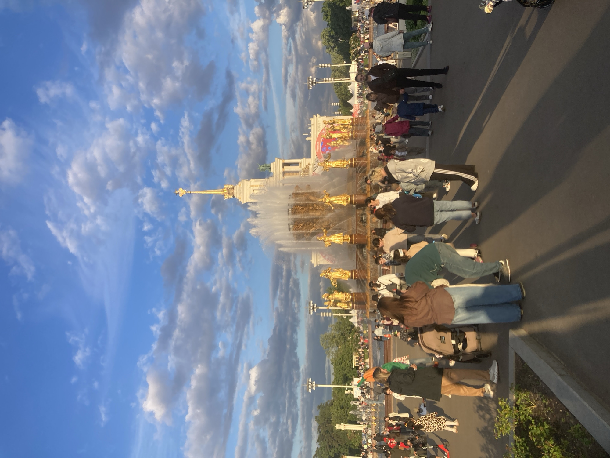 The image captures a lively outdoor scene at what appears to be a grand public park or exhibition complex, specifically identifiable as VDNKh (Exhibition of Achievements of National Economy) in Moscow, Russia. The central feature is a magnificent, multi-tiered golden fountain, known as the "Friendship of Peoples" fountain, adorned with numerous golden statues of allegorical figures and actively spouting water. Behind the fountain, a majestic, light-colored building with classical architectural elements and a prominent golden spire stands as a backdrop.
A significant crowd of people populates the wide paved area in the foreground and around the fountain. Individuals of diverse ages are seen strolling, standing, and observing the impressive fountain and building. Some people are leaning forward or crouching, possibly engaged in photography or interacting with children. A baby stroller is visible within the crowd, suggesting the presence of families enjoying the leisure environment.
The time of day appears to be late afternoon or early evening, evidenced by the warm, directional sunlight illuminating the golden structures of the fountain and casting long shadows across the paved ground. The sky is partially cloudy, showcasing patches of bright blue interspersed with soft, white, and grey clouds, indicating pleasant weather conditions. Several tall light poles are scattered throughout the grounds, suggesting the area is also active during the evening. No visible text is discernible in the image. The overall atmosphere conveys a sense of public enjoyment and sightseeing at a popular cultural attraction.