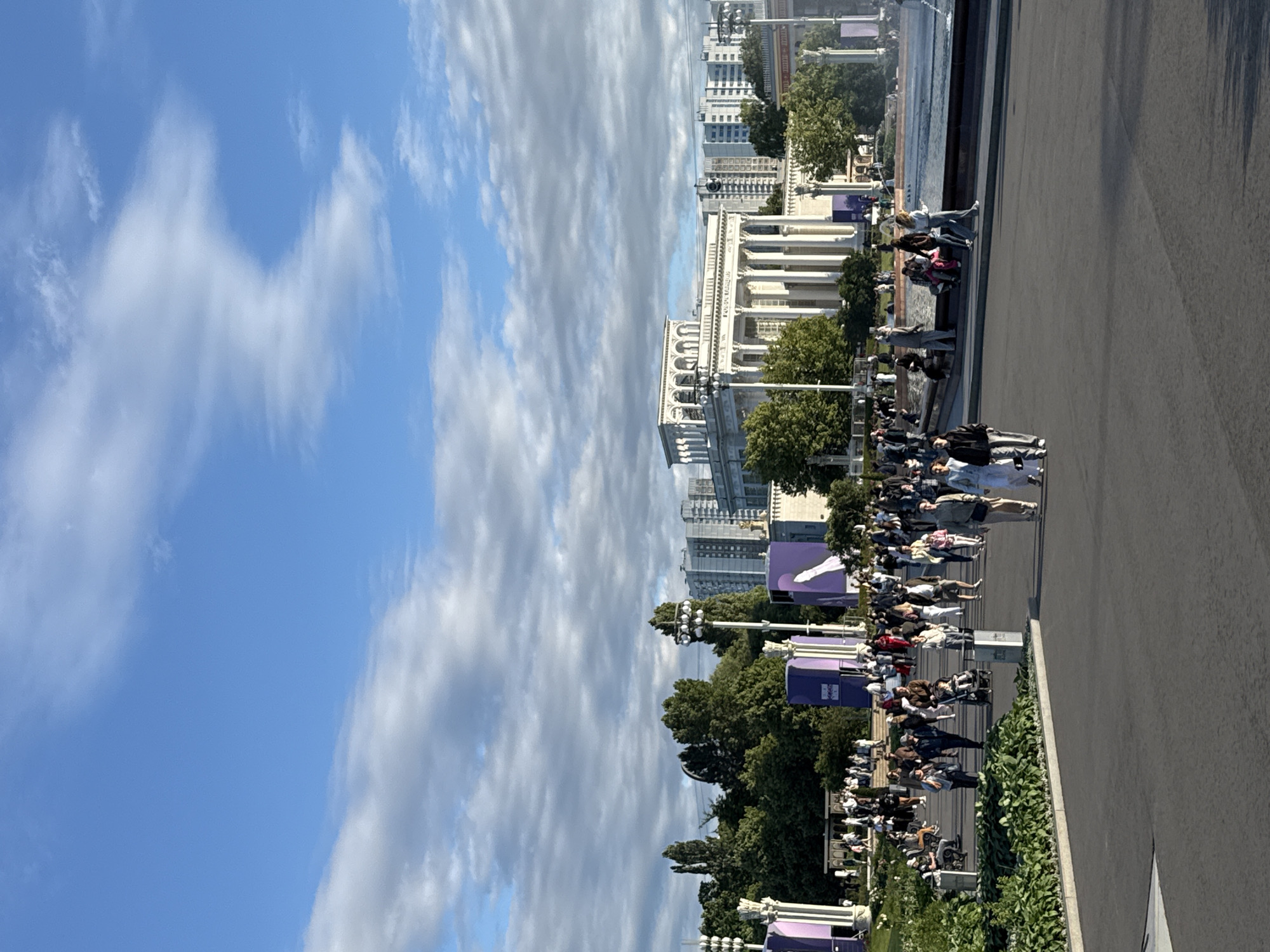 This is a photograph taken at VDNKh (Exhibition of Achievements of National Economy) in Moscow, Russia. The scene depicts a public space with many people walking and congregating. In the background, a grand white classical-style building with columns and arches is visible, likely an exhibition pavilion. Adjacent to it, tall modern residential buildings rise above the trees. The sky is blue with scattered white clouds. Several large purple structures, possibly temporary installations or exhibition booths, are scattered throughout the area. People are seen in various states of activity: walking, sitting, and pushing strollers. Some individuals are walking in groups, suggesting social interaction. The ground is paved, and there are some green spaces with trees and hedges. The overall atmosphere appears to be one of a lively public gathering on a pleasant day.