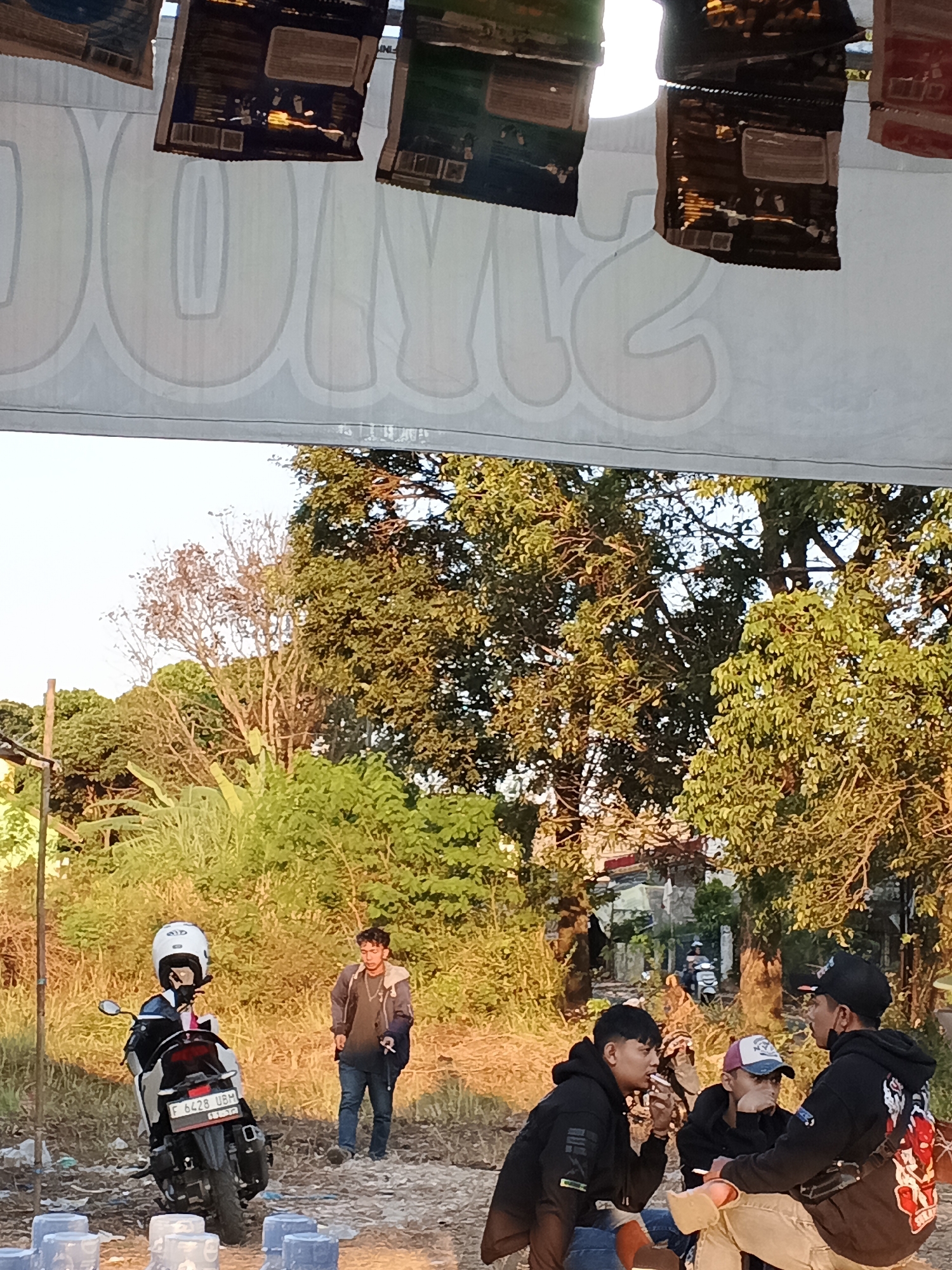 A group of young Indonesian men are gathered outdoors in a semi-rural area, possibly on the outskirts of Sukabumi, Indonesia. The scene is illuminated by the warm glow of late afternoon sunlight filtering through a canopy of lush green trees.
In the foreground, three young men are seated, engaged in conversation. Two of them wear black hooded sweatshirts, and one is smoking a cigarette. The third young man is wearing a baseball cap and light-colored pants. Their casual attire and relaxed postures suggest they are friends socializing.
To the left of the group, a motorcycle is parked. A person wearing a white helmet is seated on the motorcycle, facing away from the camera. Behind them, another young man in a dark jacket and jeans stands looking in their direction.
The environment appears to be a dusty, open area with sparse vegetation and some scattered debris, contrasting with the dense foliage of the trees in the background. A white banner with large, faded letters is stretched across the top of the frame, partially obscuring the sky. The overall impression is of an informal gathering in a relaxed, outdoor setting.