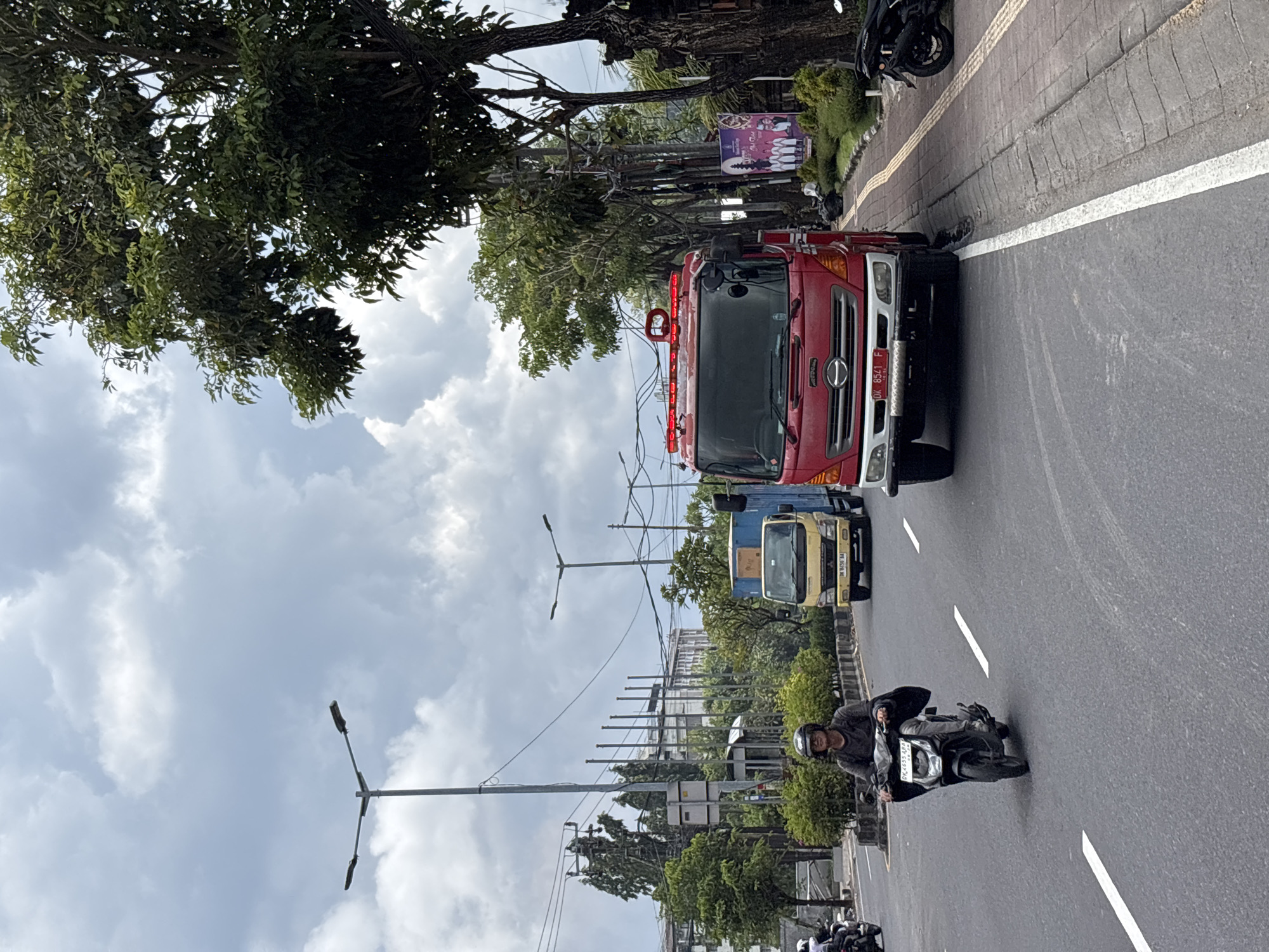 On a sunny day in Kuta, Indonesia, a fire truck is driving down a road with white lines. A motorcyclist is in front of the truck. Another truck is behind the fire truck. There are trees and buildings on either side of the road. A billboard is on the right side of the road. Streetlights are visible along the road.