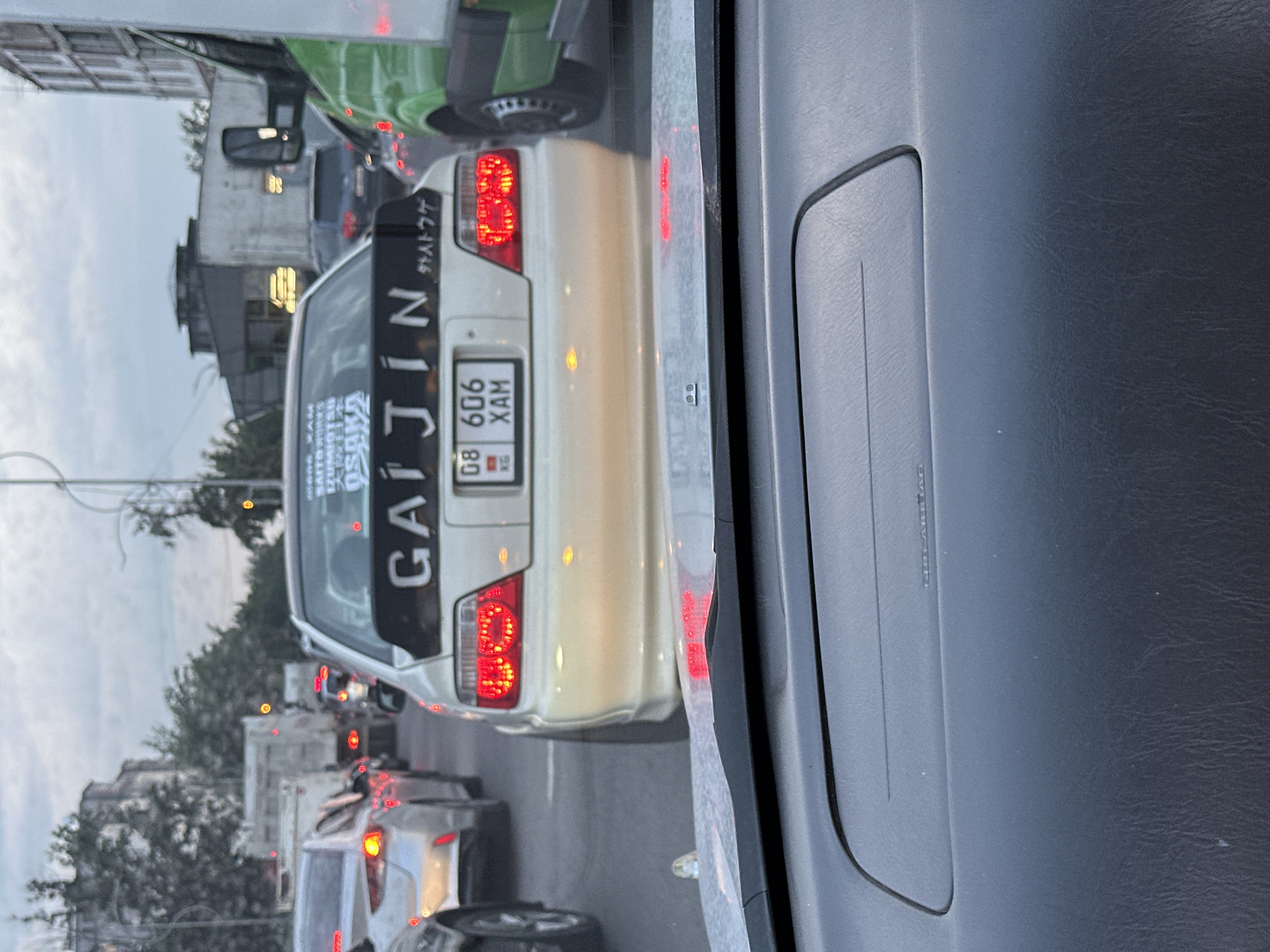 In Bishkek City, Kyrgyzstan, a beige sedan is stuck in moderate traffic on an overcast day. The car has a large black spoiler with white lettering that reads "GAI JIN" and Japanese characters. A license plate with the numbers "08 606 XAM" is visible below the spoiler. The vehicle's taillights are illuminated. The surrounding environment includes other cars, trucks, and buildings typical of an urban setting. The perspective is from inside another vehicle, looking forward through the windshield.
