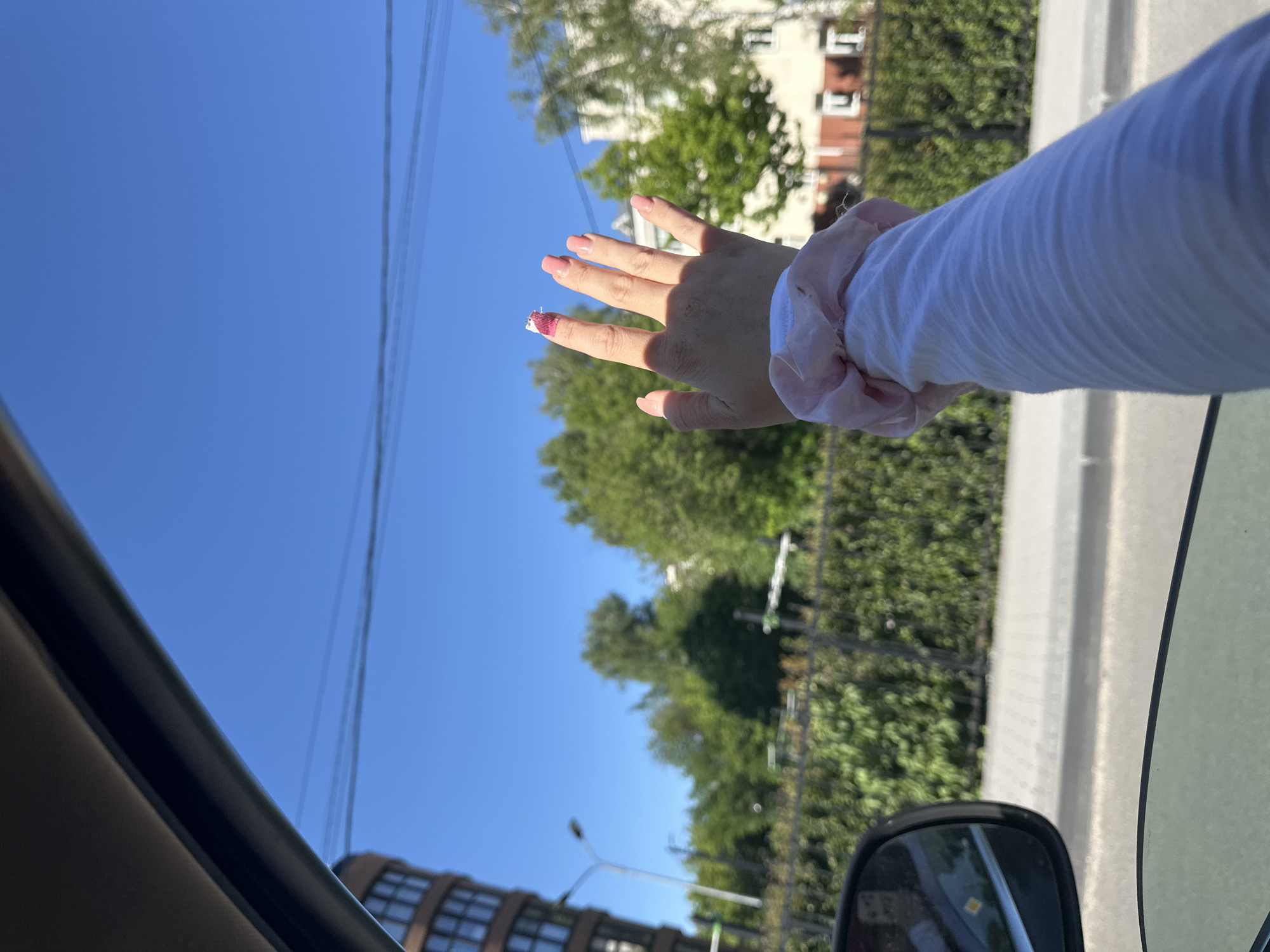 The image depicts a first-person view from inside a vehicle on a clear, sunny day in Nizhny Novgorod, Russia. A fair-skinned right hand, adorned with light pink nail polish on four visible fingers, extends out of the open car window. A small, white and pink patterned adhesive bandage is affixed to the distal phalanx of the index finger. A light pink ruffled scrunchie is worn on the wrist, which is covered by a white, long-sleeved shirt exhibiting horizontal pleats or wrinkles.
The outdoor environment features a cloudless blue sky, traversed by multiple thin, dark overhead power lines in the upper middle ground. To the far left, a distinctive, multi-story residential building with a curved, reddish-brown facade and numerous square windows is prominent. This building is specifically identified as the residential complex located at **Proviantskaya Street, 7k1, Nizhny Novgorod**. To the right of this building, a tall, white street light pole stands. A dense line of green trees and hedges forms a barrier along the roadside, behind which additional green trees and a white building with red window frames are visible further to the right. A paved road and the edge of a sidewalk are visible in the lower portion of the exterior scene. The dark interior frame of the car window is visible at the top, and a portion of the car's side mirror, reflecting a yellow diamond-shaped road sign, is present in the lower left corner. No other individuals are visible in the frame.