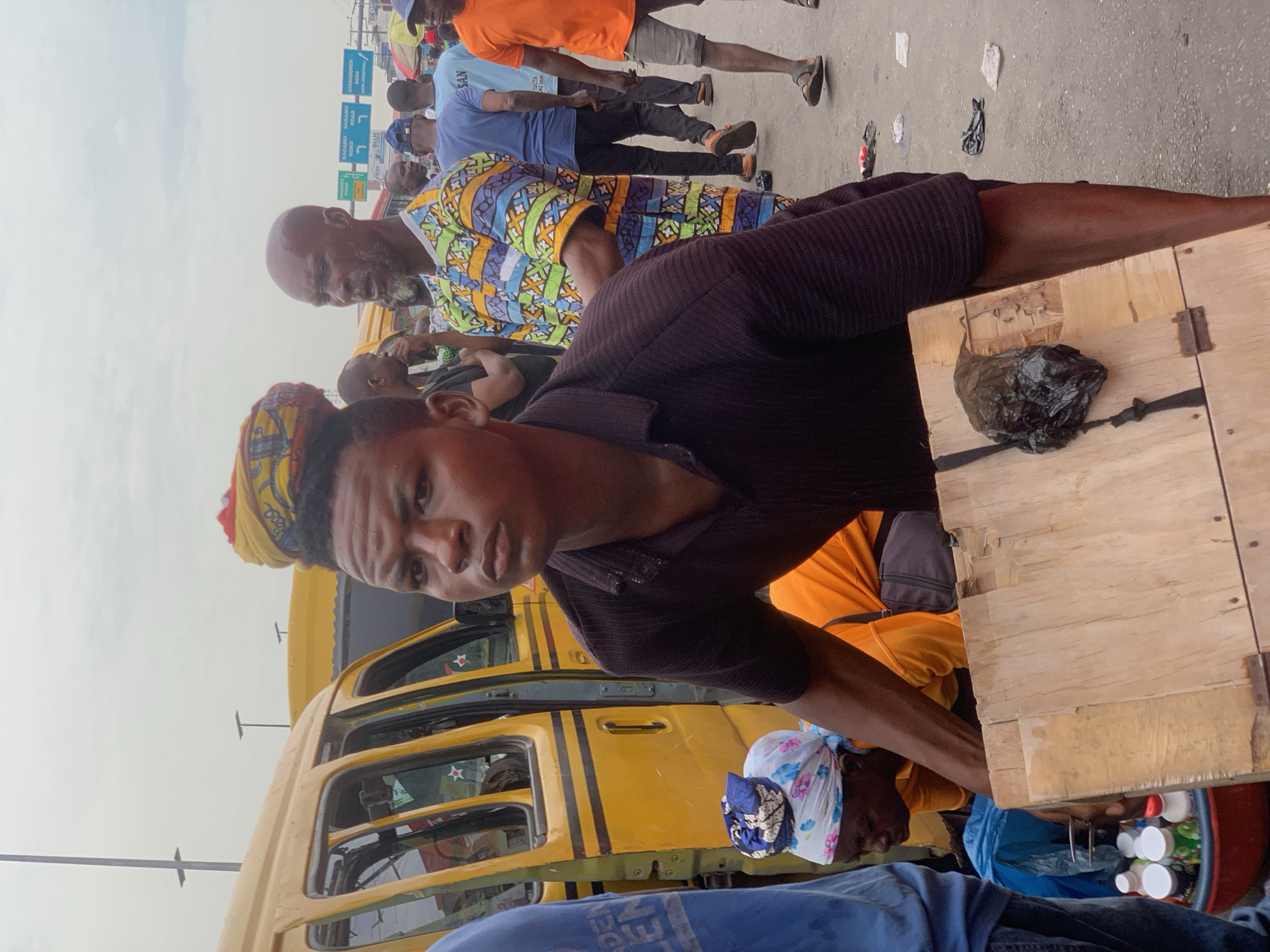 The image captures a bustling scene in Ikorodu, Nigeria. A young man in a dark brown shirt and a yellow and red headwrap stands prominently in the foreground next to a wooden food cart. Behind him is a yellow "danfo" bus, a common mode of transportation in Lagos. Other people are visible in the background, including one in a patterned shirt. The wooden food cart has a black plastic bag of goods placed on top. Underneath the cart, plastic containers and a blue bag are visible. In the distant background, signage indicates directions to Ayangburen Road and Sagamu Road. The overall atmosphere appears to be that of a busy, crowded market street.