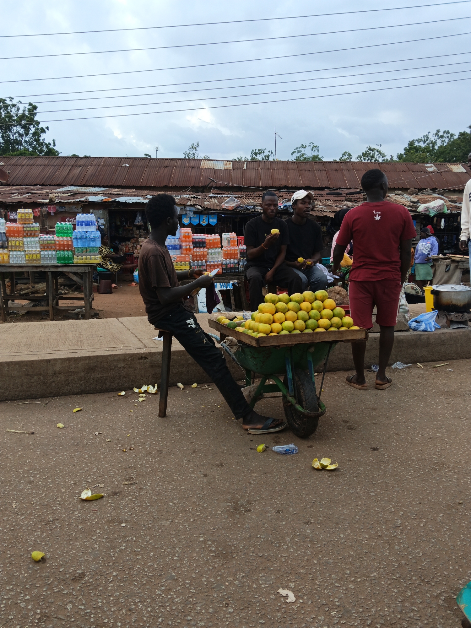 The image depicts a bustling outdoor market scene in Bukuru, Nigeria. Several individuals are present, some appearing to be vendors and others customers or bystanders. In the foreground, a man is seated on a stool next to a wheelbarrow overflowing with a large quantity of oranges. He is casually holding a piece of fruit and looking to the side. Behind him, three other men are standing and talking, with two of them holding fruit.
The market setting is characterized by makeshift stalls with corrugated metal roofs, suggesting a somewhat rustic or informal marketplace. Stacks of bottled drinks, likely water and soft drinks, are visible on tables and shelves behind the vendors. The ground is paved with asphalt, and there are scattered pieces of discarded fruit peels.
The sky is overcast with clouds, indicating a potentially cloudy or overcast day. The overall atmosphere appears to be that of daily commerce and social interaction in a busy Nigerian market. There are no explicit timestamps, but the lighting suggests it is daytime, possibly late morning or afternoon.
Visible text includes "Coca-Cola" on a bottle stack and some indecipherable text on the clothing of some individuals. One man wears a maroon t-shirt with a white emblem resembling a leaf.