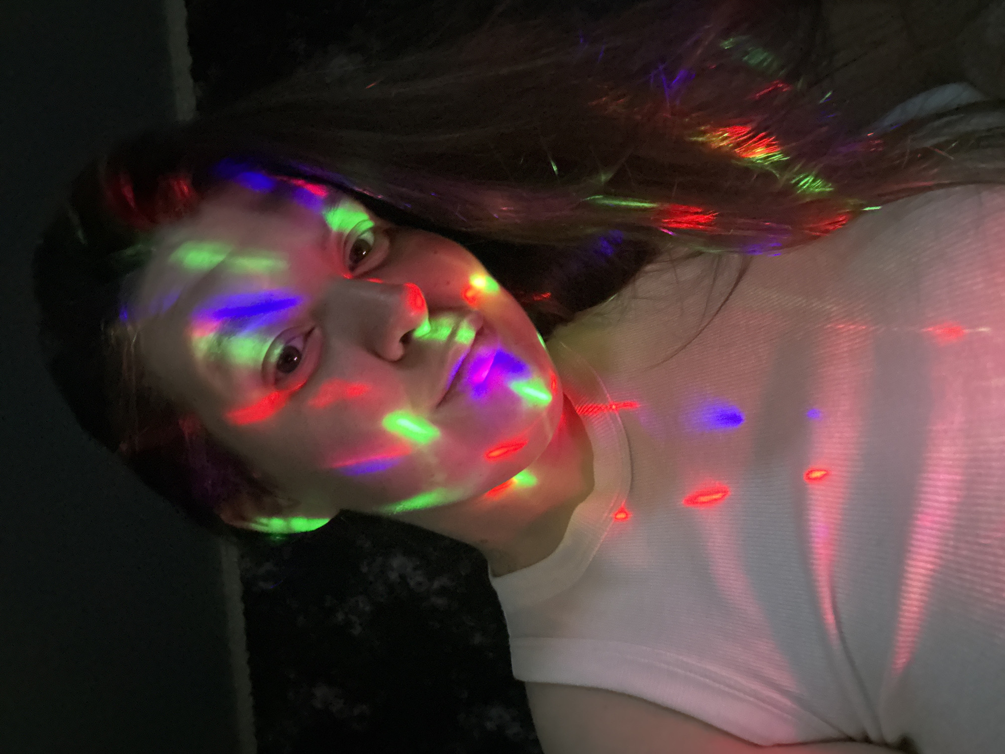 A woman is pictured indoors under multicolored light. The lights are red, green, and blue and appear to be projected onto her face and white shirt. The background is dark and indistinct. The photo appears to be taken at close range. The image's location is unknown.