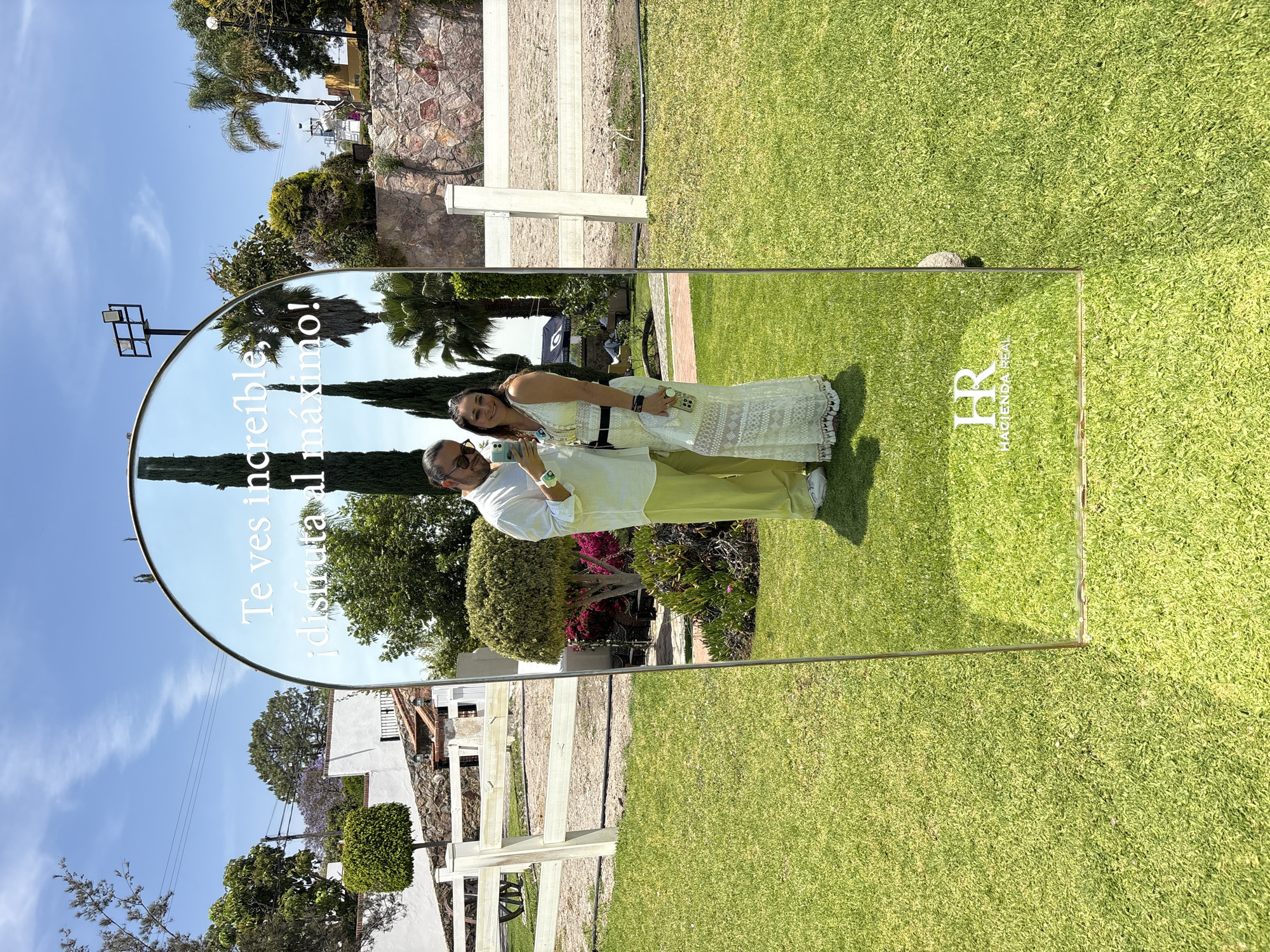 The image captures a couple reflected in a mirror outdoors in El Oasis Valsequillo, Mexico. The couple is standing on a well-manicured lawn. The man is wearing a white shirt and light green pants, while the woman is in a white, lace dress. They are holding phones and taking a photo of themselves in the mirror. The mirror has a curved top and the phrase "Te ves increible ¡disfruta al máximo!" written on it. The logo "HR HACIENDA REAL" is visible in the lower right corner of the mirror. The reflection in the mirror reveals a backdrop of trees, a white fence, and buildings. The environment is bright and sunny.