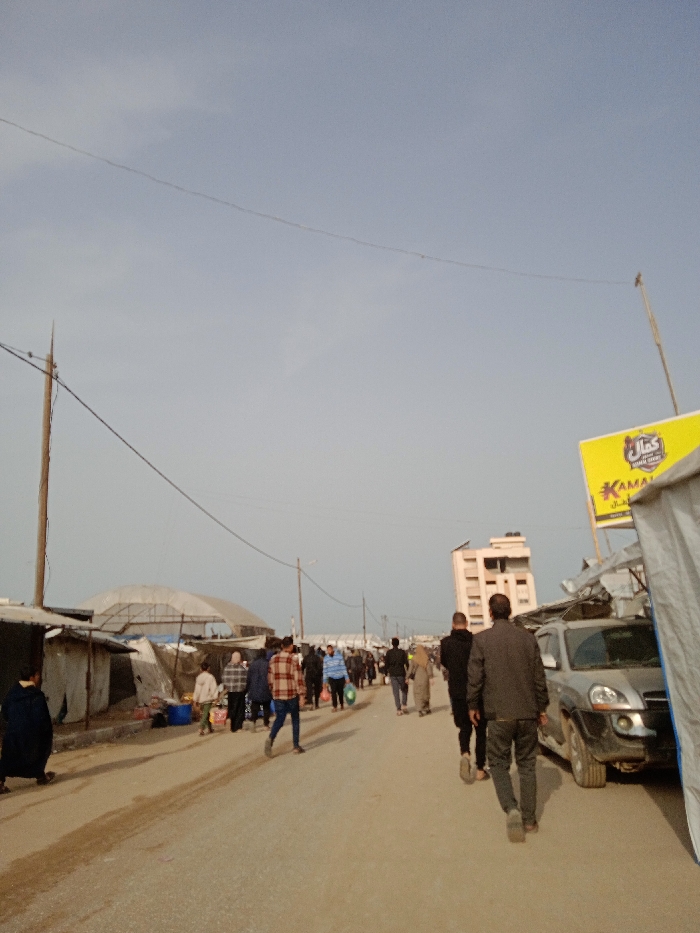 The image depicts a crowded, unpaved street or thoroughfare in Khan Yunis, Palestinian Territory, under a clear, light blue sky. In the foreground on the left, a person in dark clothing is crouched beside makeshift shelters constructed from corrugated metal and tarps. These rudimentary structures extend along the left side of the road, featuring a prominent, large white arched canopy further back. A wooden utility pole with multiple electrical wires crosses the upper left frame. On the right, a silver SUV, possibly a Hyundai Tucson, is parked, partially obscured by a large, light-colored canvas or tarp structure. Above this, a bright yellow rectangular sign with Arabic text, including "كمال" (Kamal), is visible. Numerous individuals, predominantly adults, walk down the center of the road away from the viewer. Their attire varies, including plaid shirts, dark jackets, and headscarves for some women; some carry bags. In the background, a multi-story building with a light-colored facade is visible between the walking crowd and the right-side structures. Another utility pole stands further down the road on the right.