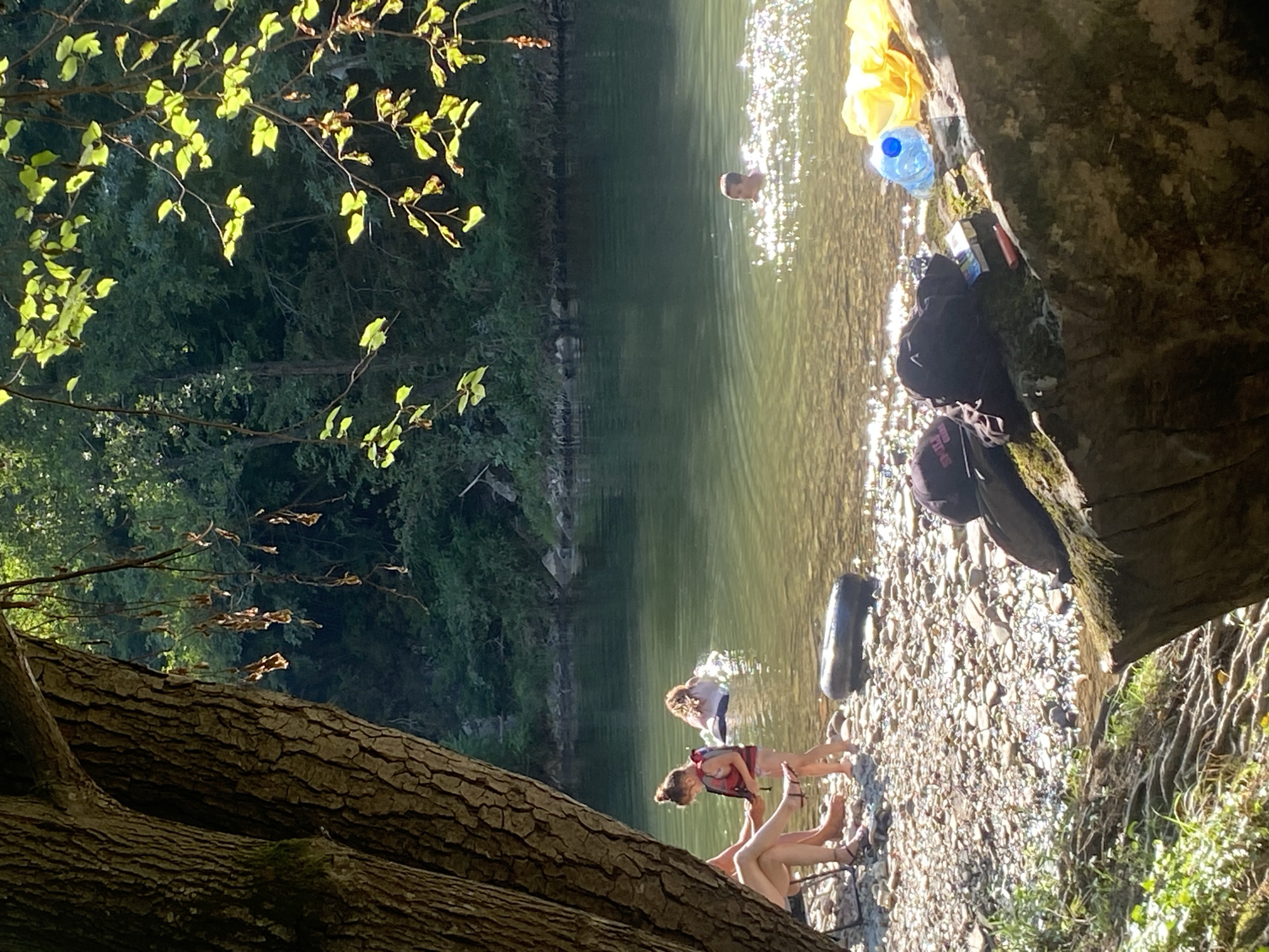 The image depicts a sunny day at a riverbank, likely in or near the town of Debnevo, Bulgaria. The foreground shows a rocky shore with various items: a large tree trunk on the left, a black inner tube, and on a large rock to the right, several objects are placed, including two backpacks, a pack of cigarettes, a lighter, and a blue plastic water bottle. Two individuals are seated in a camping chair on the left, their legs visible. A young girl in a life vest stands near the water's edge. Further in the river, another person is swimming, and a third individual is partially visible in the water further out. The river itself is calm with sunlight reflecting off its surface. The background consists of dense green trees and foliage lining the opposite bank of the river.