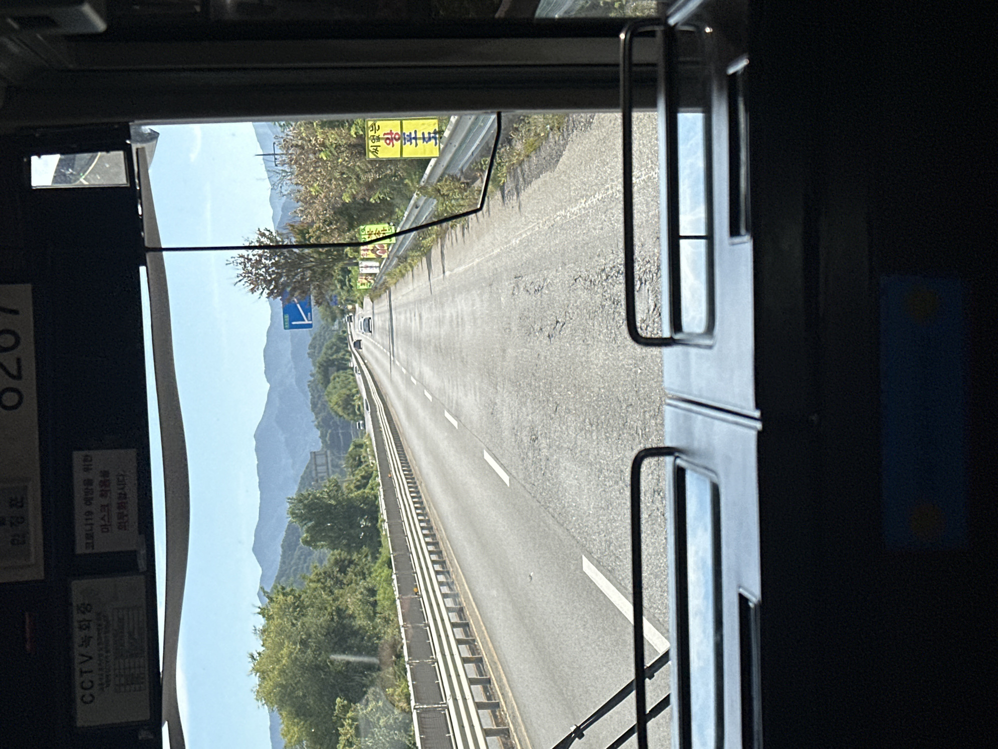 The image is taken from the interior of a bus, looking out through the windshield. The bus appears to be traveling on a highway, indicated by the multi-lane road and guardrails. On the right side of the road, there are several yellow signs with Korean text. In the distance, mountains are visible under a clear blue sky. On the interior of the bus, above the windshield, there are two signs. One has the text "CCTV 녹화중" which translates to "CCTV Recording". The other sign reads "코로나19 예방을 위한 마스크 착용을 의무화합니다" which translates to "Wearing a mask is mandatory for COVID-19 prevention". The dashboard of the bus is visible in the foreground, with two metallic bars. The overall impression is a scenic drive on a highway in South Korea, with visible public service announcements related to CCTV and COVID-19 prevention. Without further information, the exact location in Jeonju-si cannot be determined from the visual data alone, but it is clearly a road in South Korea.