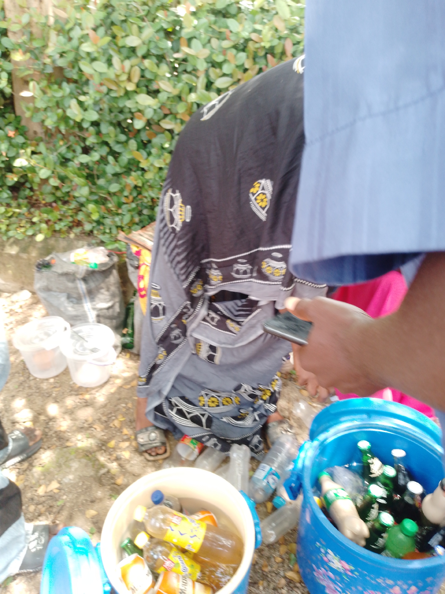 The image depicts an outdoor scene with lush green foliage in the background. In the foreground, a person is holding a smartphone. Scattered around are several plastic bottles and buckets, some filled with beverages. A large black bag, possibly a trash bag, is also visible. The ground is covered with dirt and fallen leaves. The overall impression is that of a casual outdoor gathering or perhaps a cleanup effort. The specific location is in Akwanga, Nigeria.
