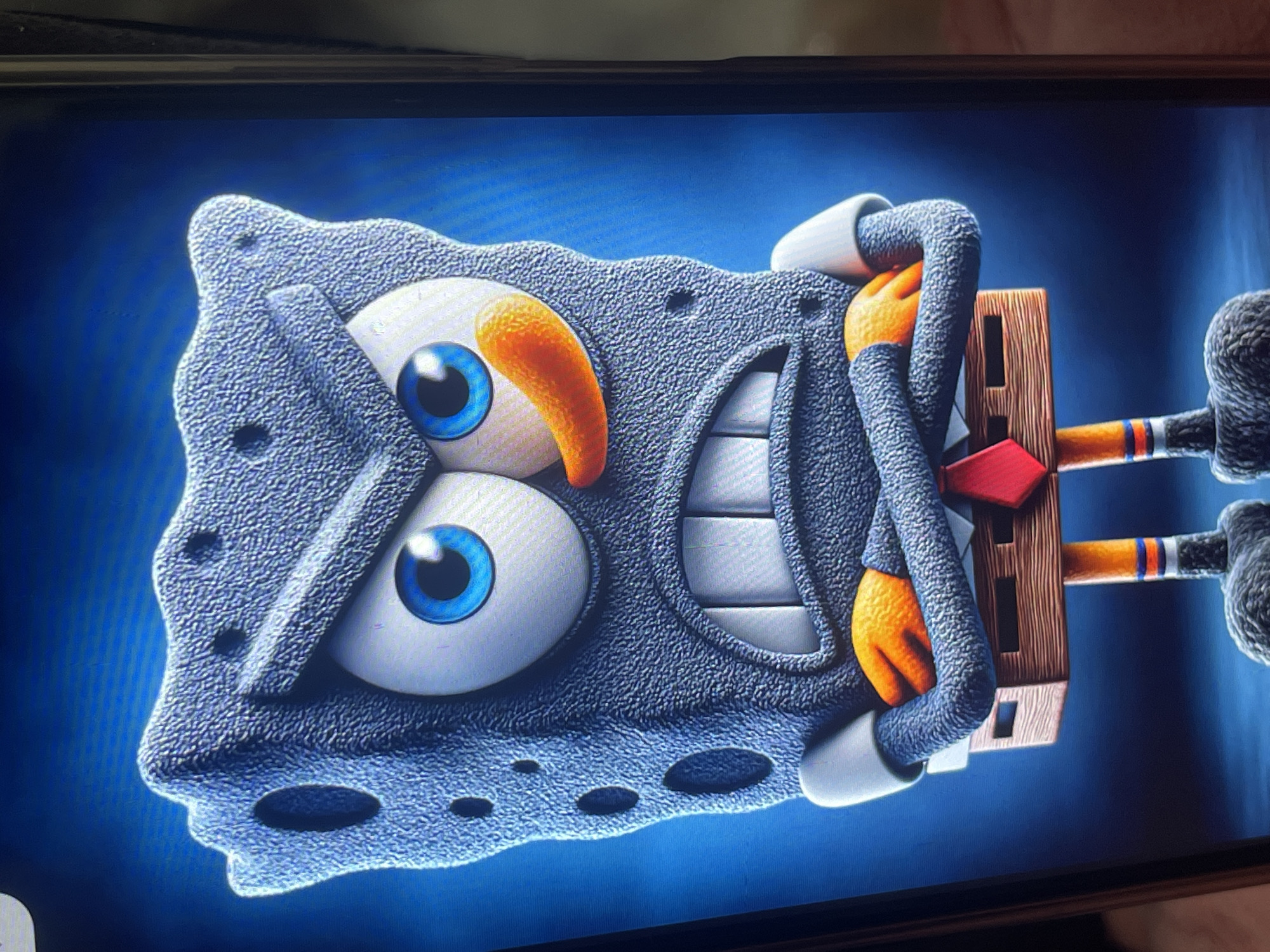 The image shows a close-up of a stylized, anthropomorphic sponge character, resembling SpongeBob SquarePants, rendered in a textured, grey material. The character has large, expressive blue eyes with thick white rims, an orange, pointed nose, and a wide, gritted-teeth smile revealing white teeth. It is wearing a white collared shirt with a red tie, and its arms are crossed over its chest with orange, five-fingered hands. The lower body of the character is composed of wooden-like planks, with orange legs featuring blue and white striped socks and grey, furry feet. The background is a dark, gradient blue, suggesting a studio or digital environment, with no discernible elements of Bishkek City, Kyrgyzstan.