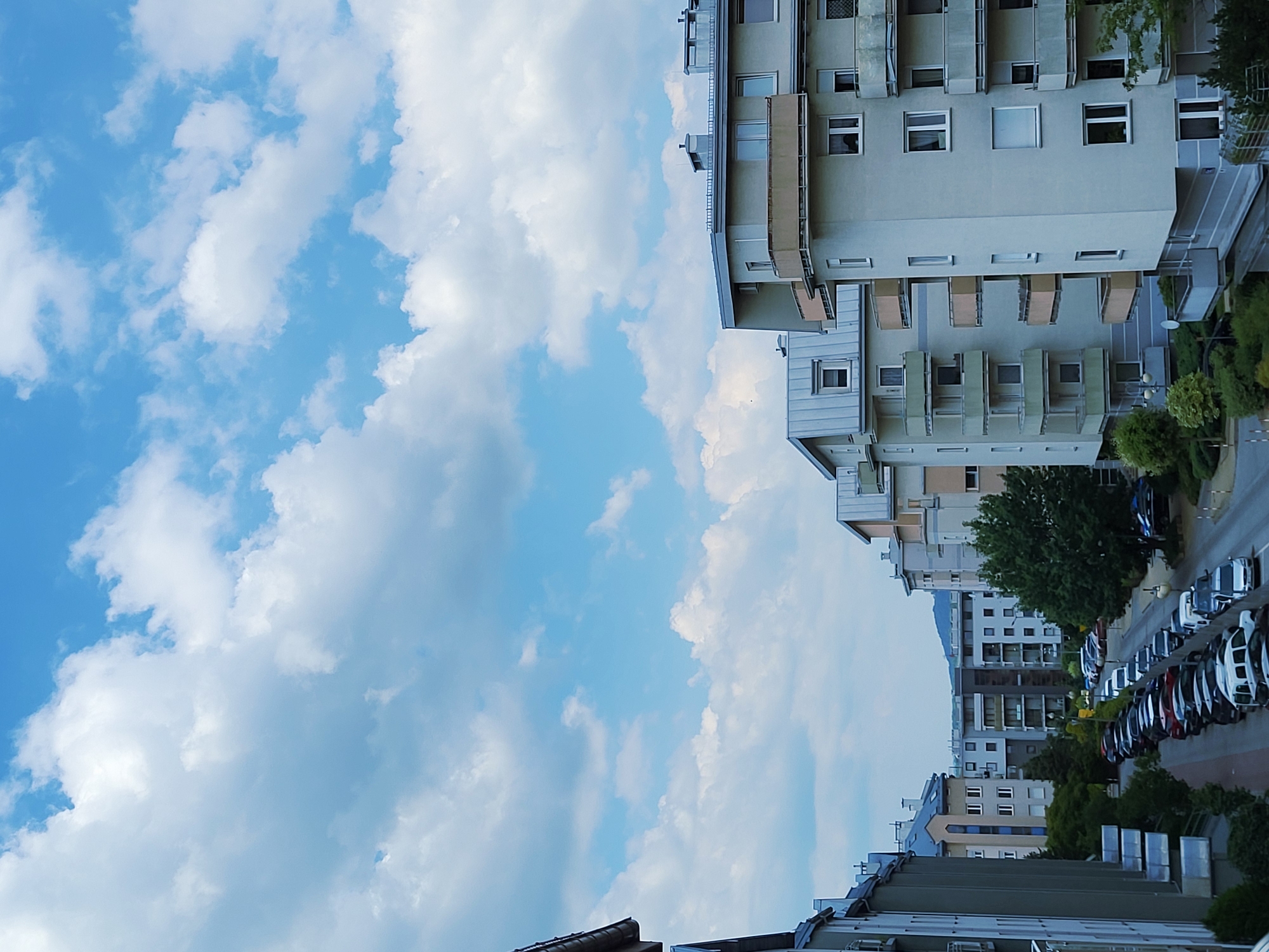 The image shows a street lined with apartment buildings and parked cars, under a bright, cloudy sky. The buildings are modern, with balconies and large windows. The street appears to be in a residential area, with trees and green spaces visible. Several cars are parked along the street, including sedans, SUVs, and vans. The sky is blue with scattered white clouds, indicating good weather. The scene is well-lit, with no obvious signs of activity or people.