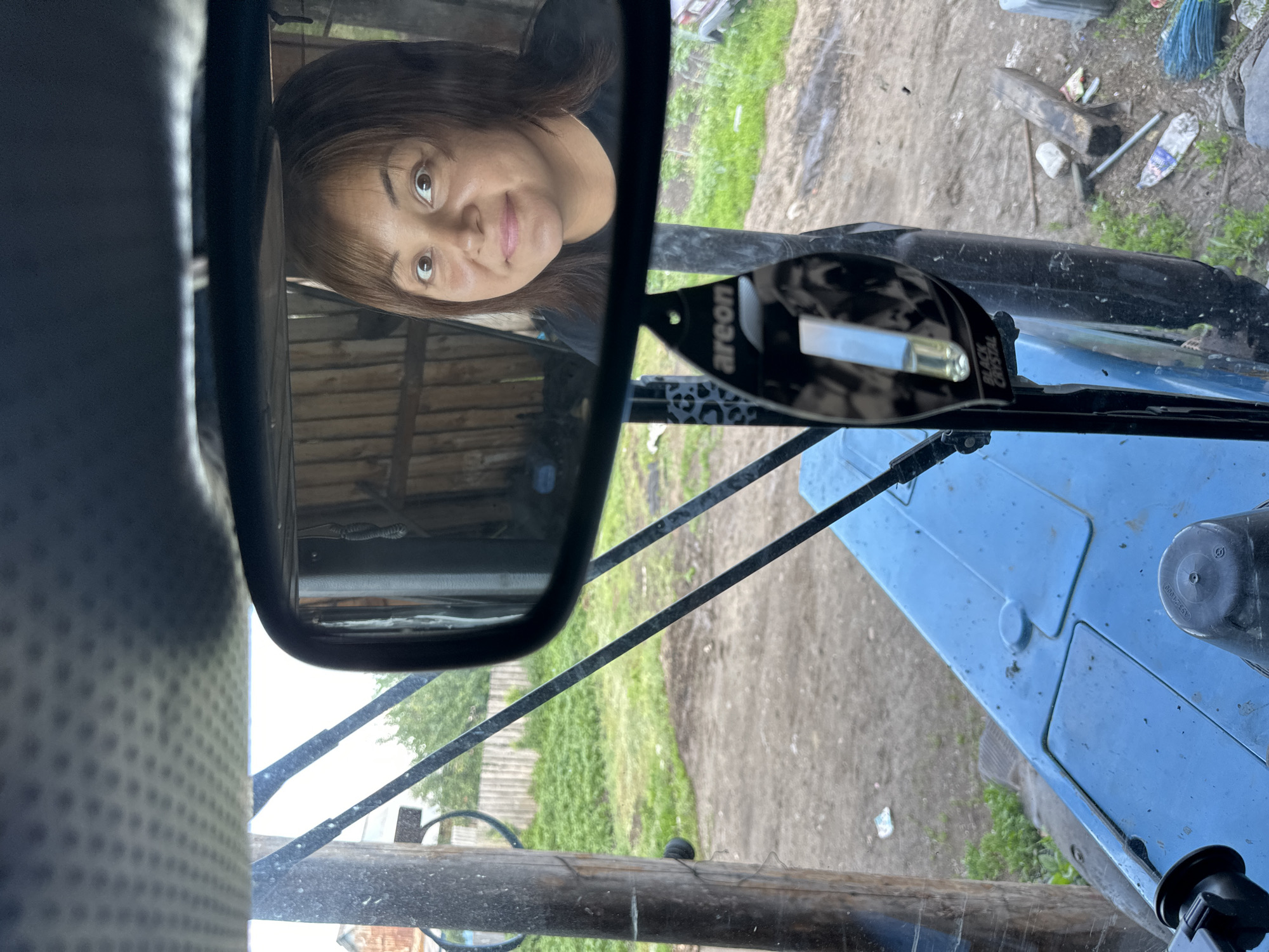 The image is taken from the perspective of someone inside a blue tractor. A rearview mirror reflects the face of a woman with brown hair and brown eyes, wearing a black t-shirt. She is smiling. In the foreground, part of the tractor's blue interior is visible, including a steering wheel and control panel. A black air freshener with the brand "areon" is hanging from the mirror. Outside the tractor, a wooden fence and green foliage can be seen, suggesting an outdoor, possibly rural setting. There are also some tools and debris scattered on the ground.