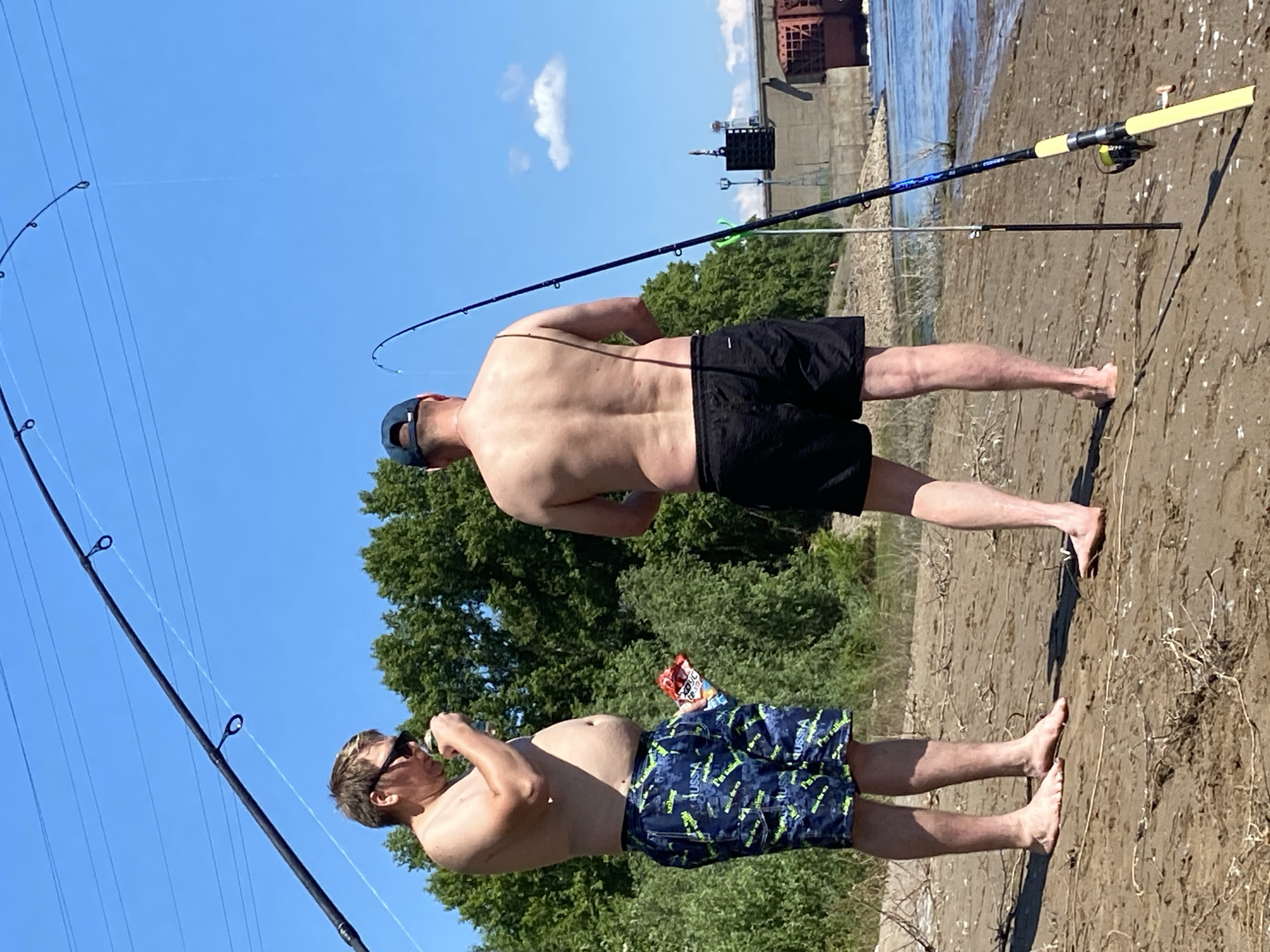 Two men are fishing on the sandy bank of the Kama River in Chaikovsky, Russia. The man on the left is wearing patterned swim trunks and sunglasses and is holding a can and a bag of snacks. The man on the right is wearing black swim trunks and a blue baseball cap. Both men are shirtless and standing with their fishing rods cast into the water. In the background, there are trees, a dam structure, and power lines stretching across the clear blue sky.