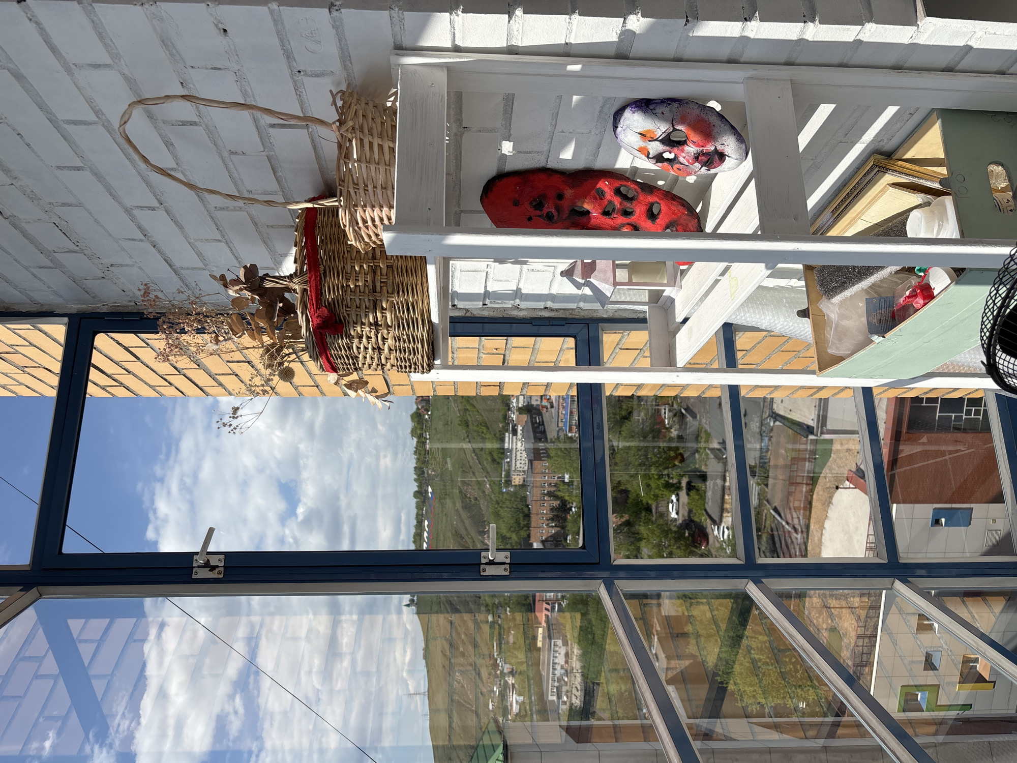 The image depicts an interior space with a view through a window. The view shows a landscape with green hills and buildings, and reflections of the sky on glass panels. A basket with dried flowers is on the left. A white shelf unit holds a red mask, a purplish mask, a picture frame, and packing materials. The brickwork is white. The building appears to be in Krasnoyarsk, Russia, based on the surrounding architecture and landscape.