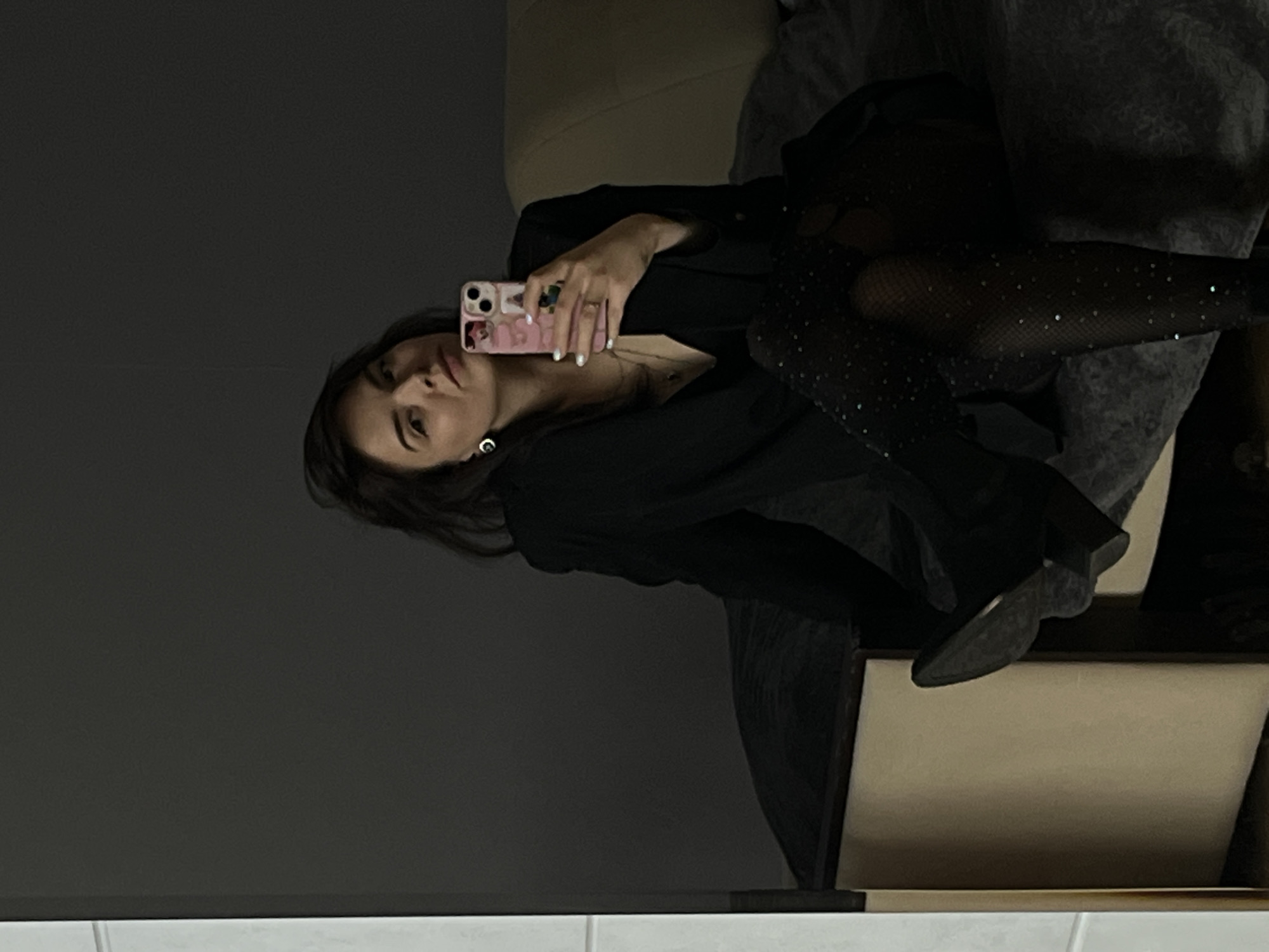 This is a mirror selfie, presented sideways, featuring a young adult female indoors. She has dark hair and is wearing a dark, possibly V-neck, top or dress. A prominent feature of her attire is a pair of black fishnet stockings adorned with small, shimmering rhinestones, and she is wearing dark shoes.
She is holding a pink smartphone with a decorated case in her right hand, positioned to capture her reflection. The phone case has several small charms or stickers, one of which faintly resembles the letter "C" or a similar emblem. The phone itself shows a visible "8" near its camera module, indicative of an iPhone. Her expression is neutral, with her gaze directed towards the phone's camera. She is also wearing visible earrings.
The background is dimly lit and mostly dark, suggesting an indoor setting during evening or night. Behind the woman, a light beige-colored surface, possibly a sofa or bed, is partially visible. The bottom edge of the image shows a narrow white line, hinting at a floor or another surface. The overall atmosphere is casual, documenting a self-portrait moment. This media file is associated with Shushary, Russia.