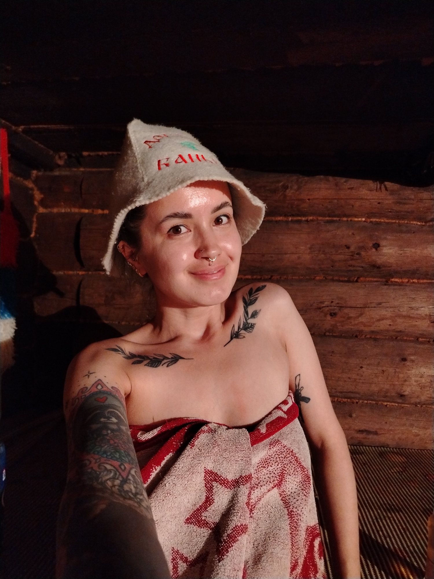 A woman is indoors, likely in a sauna with wood-paneled walls. She wears a white sauna hat with the name "KAIJU" stitched in red and green. She is wrapped in a red and white towel. She has several tattoos on her upper body and arms, including one of a raccoon on her left arm and a leaf sprig on her left shoulder. The scene is dimly lit. The city of Гожан, Russia is not apparent in the image.