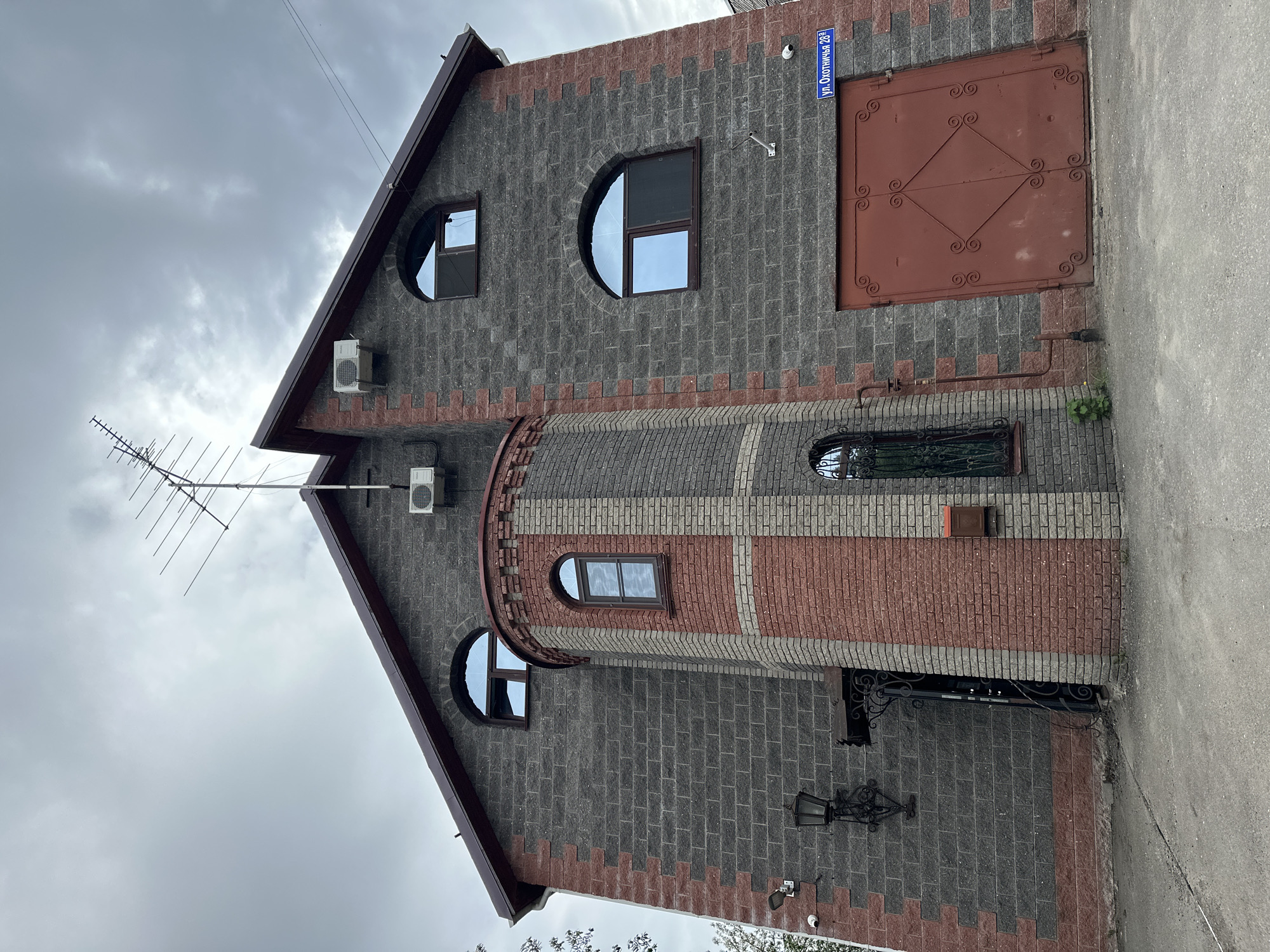 The image shows a building in Nizhny Novgorod, Russia, identifiable by the street sign reading "ул. Охотничья 23а" (Okhotnichya Street 23a). The building has a distinctive architectural style, featuring a combination of grey and red brickwork. There is a tower-like structure with alternating grey and red brick bands. The building has round windows and rectangular windows with decorative ironwork. An antenna is visible on the roof. An air conditioning unit is mounted on the wall. A large red garage door is located on the right side of the building. A street light is mounted on the brick wall near the entrance. The sky is overcast. A section of pavement or road is visible on the right side.