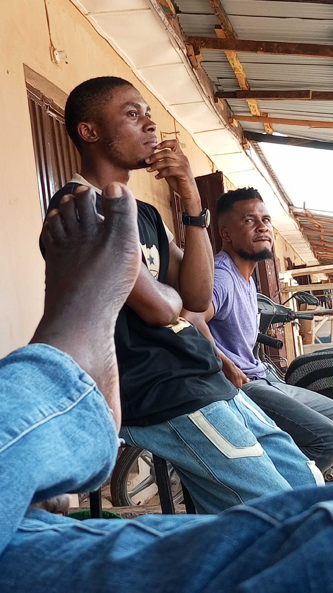 The image depicts three men seated outdoors. The man in the foreground is wearing blue jeans and his foot is prominently displayed. A second man is visible wearing a black shirt, blue jeans, and a wristwatch; he is seated with his arms crossed and his hand raised to his chin. The third man, seated further back, is wearing a purple shirt and grey pants. A motorcycle is parked nearby. The location is Sabon Tasha, Nigeria.