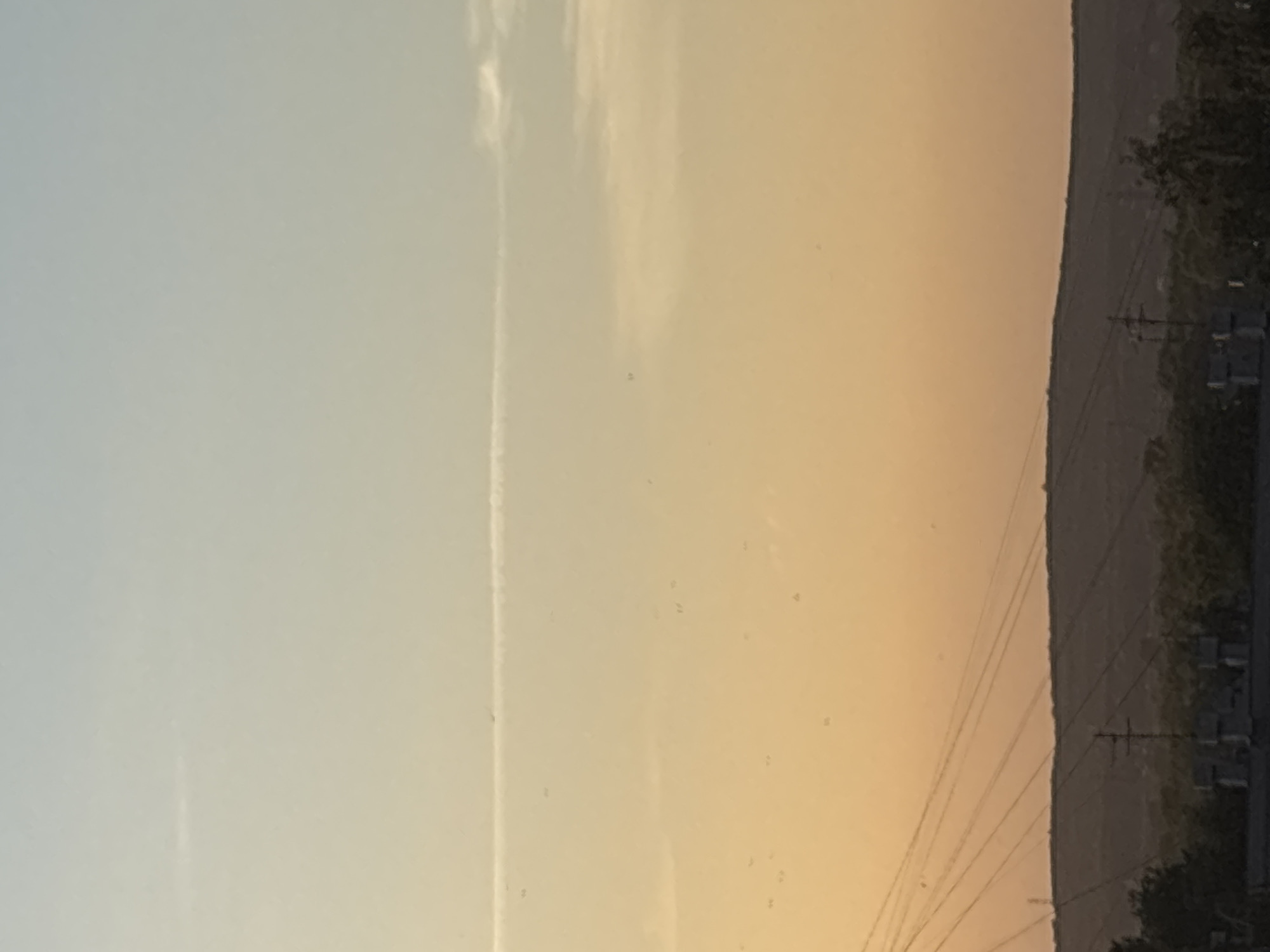 The image depicts a hazy sky with a gradient from pale blue to orange. A thin contrail cuts vertically through the sky on the left side. On the right, a dark, elevated landmass is visible, with power lines running down its edge, suggesting a city or town at its base. The precise location within the city of Unknown, Unknown, is not determinable from this image.