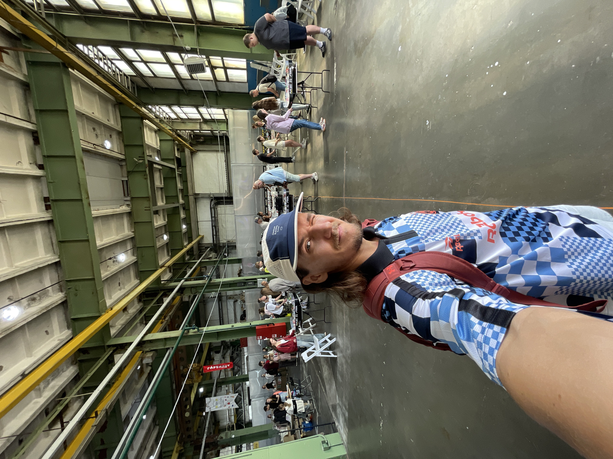 The image depicts a man taking a selfie in a large, industrial-style indoor space. He is wearing a checkered shirt and a baseball cap, with a backpack slung over his shoulder. In the background, there are several people standing around tables, some of whom appear to be playing a game. The environment is characterized by high ceilings with exposed beams and windows, suggesting it might be a former factory or warehouse. There are also banners and industrial equipment visible. The overall scene suggests a public event or gathering is taking place. The man in the foreground appears to be winking at the camera. The flooring is a polished concrete surface.